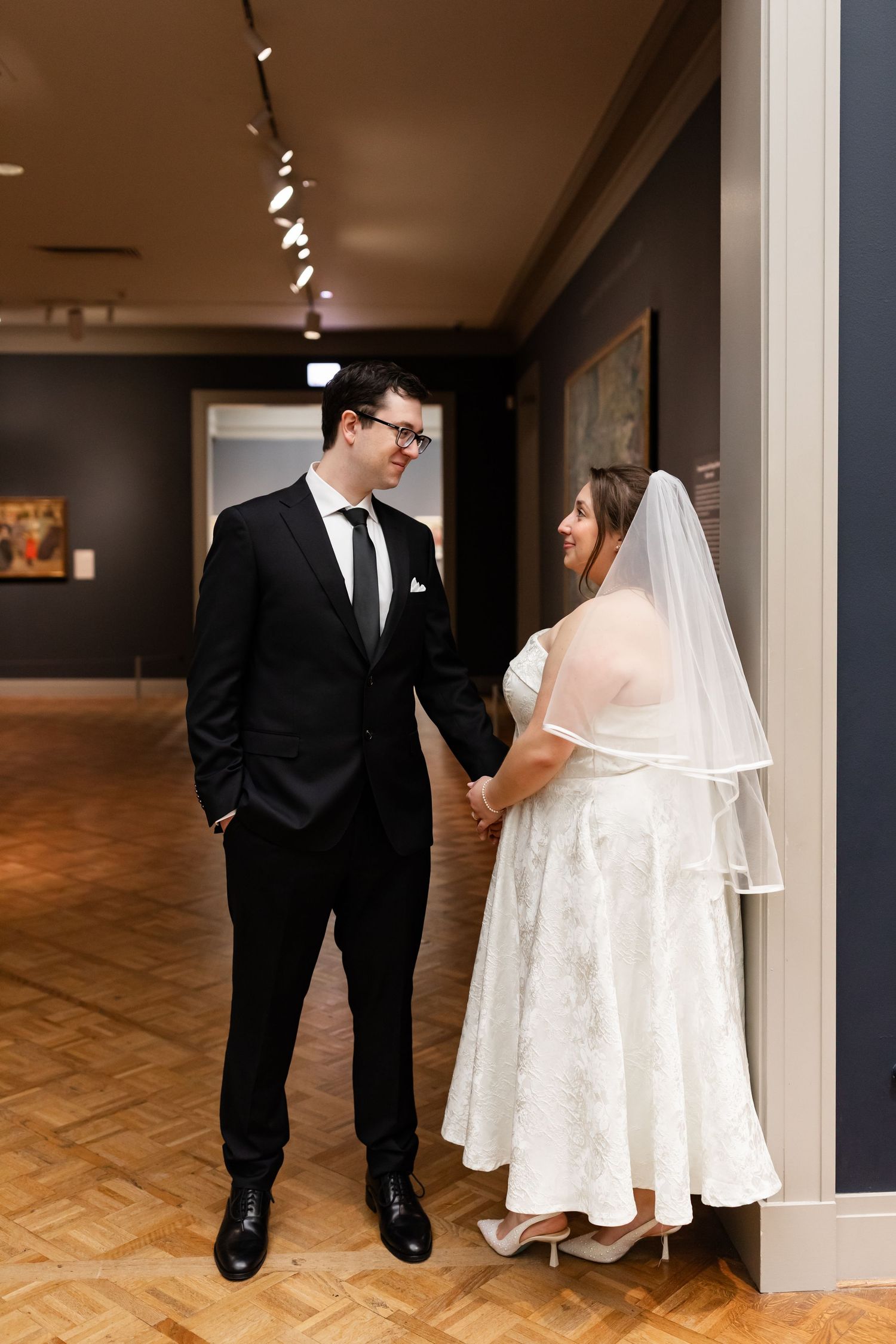 Bride and groom holding hands and smiling at each other in an elegant art museum hallway.