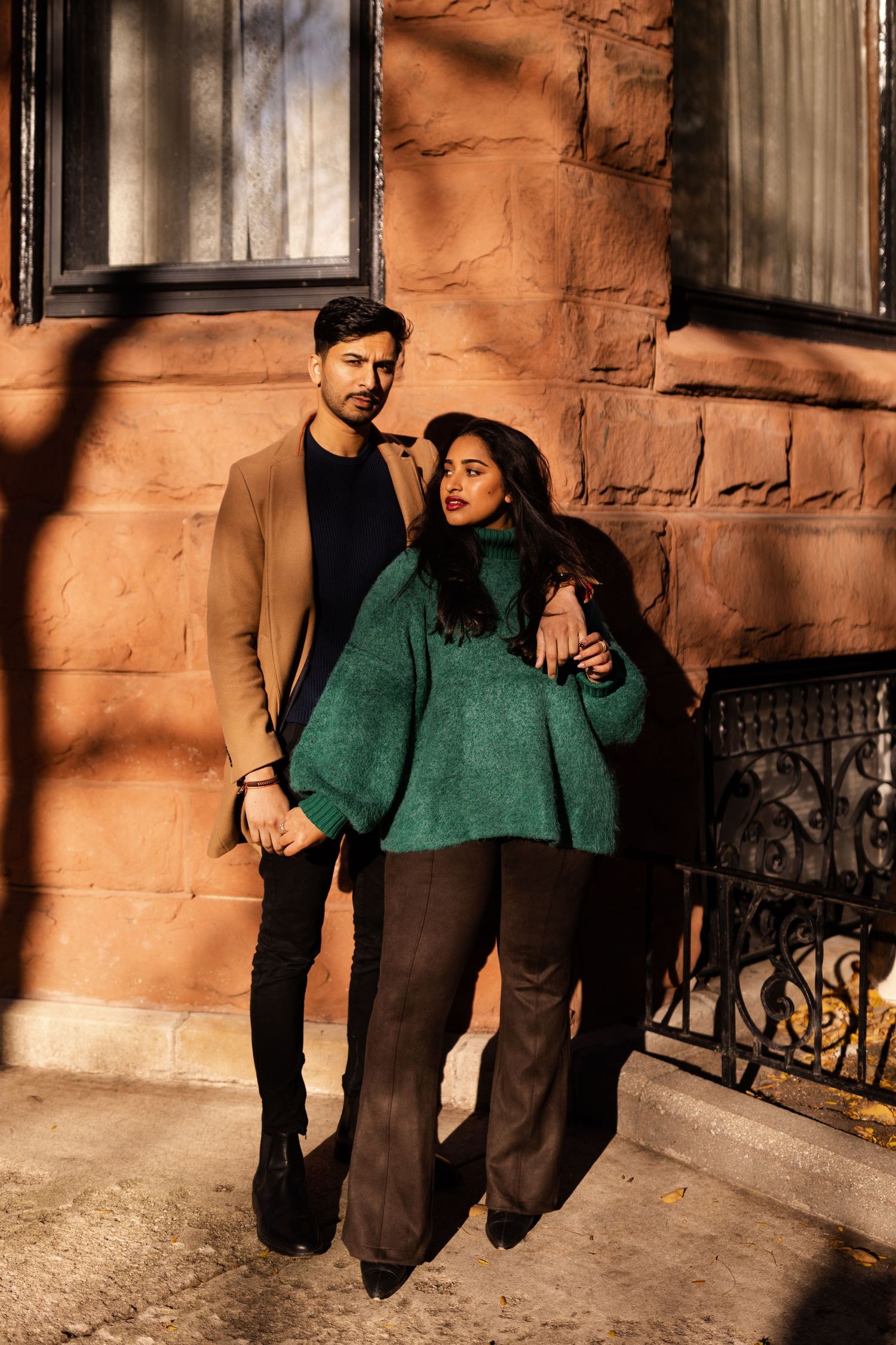 Stylish couple posing against rustic stone wall, man in camel coat, woman in teal oversized sweater and wide-leg pants.