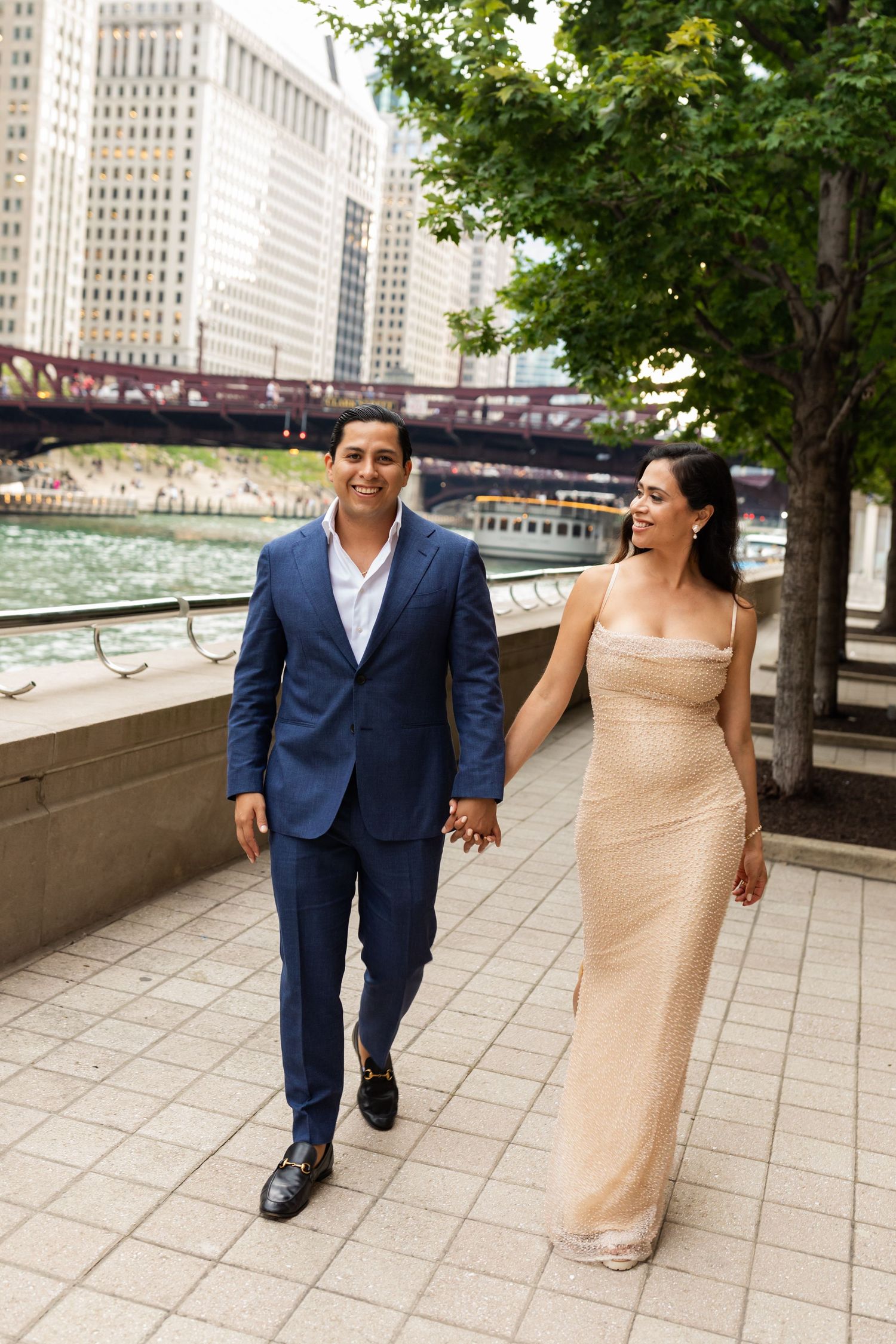 Smiling bride and groom walking hand in hand along Chicago Riverwalk, she in gold gown, he in blue suit.