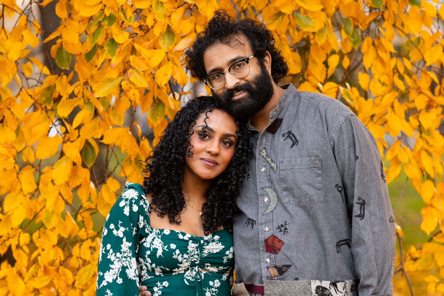 A couple smiles together in front of vibrant golden autumn leaves in an outdoor fall portrait.