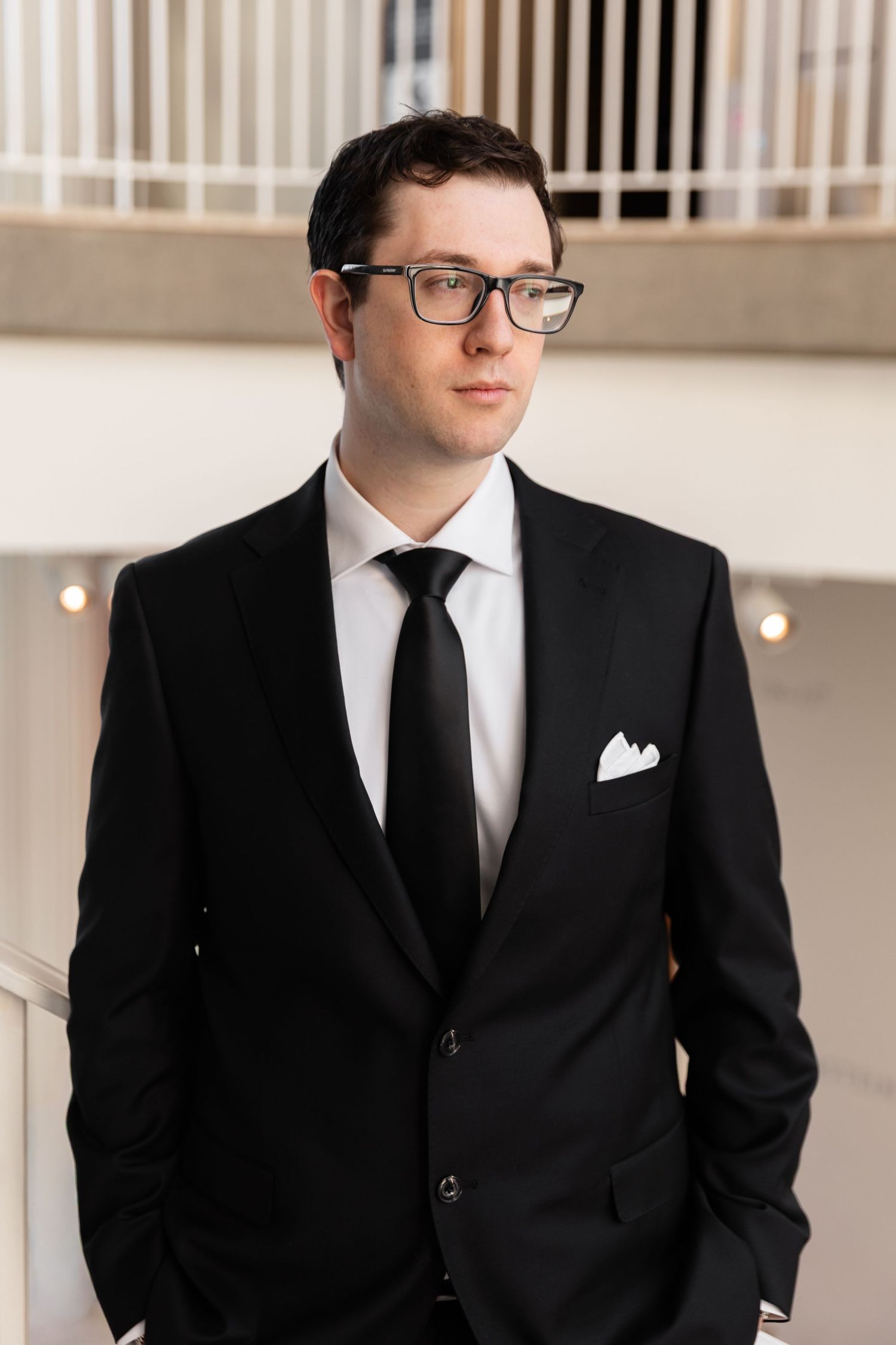 Serious groom in black suit with white pocket square and glasses standing in bright modern interior.