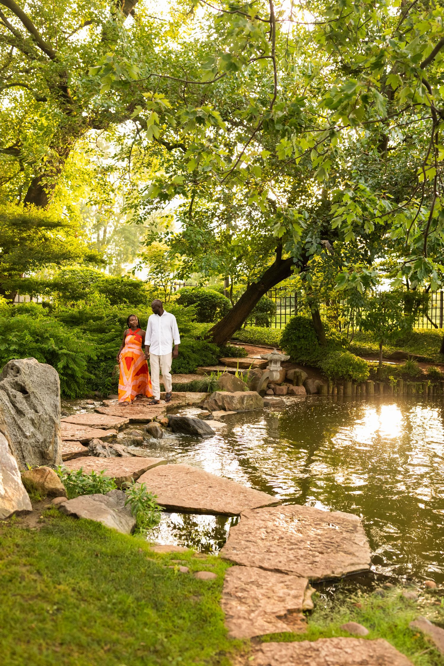 A couple walks hand in hand along stepping stones beside a serene pond in a lush Japanese garden.