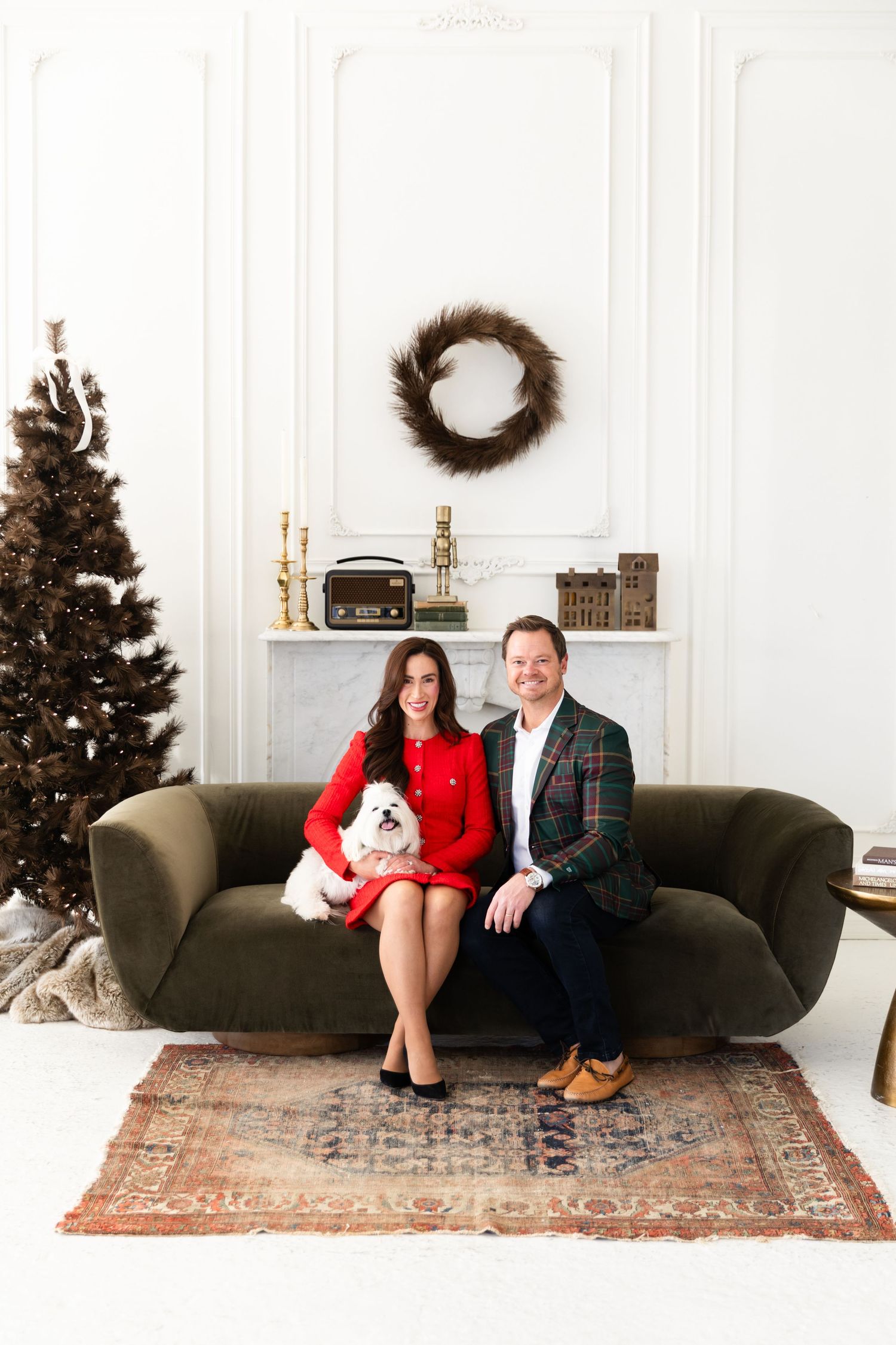 Couple in holiday attire sit on green velvet sofa with small white dog near Christmas tree and fireplace.