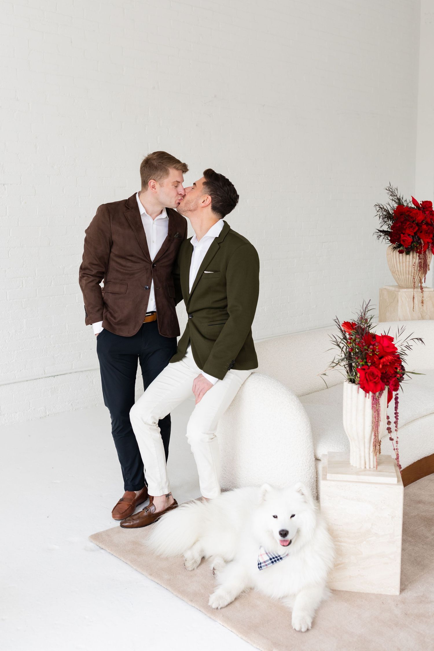 Two grooms sharing a kiss beside white Samoyed dog wearing plaid bandana with red floral decor backdrop.