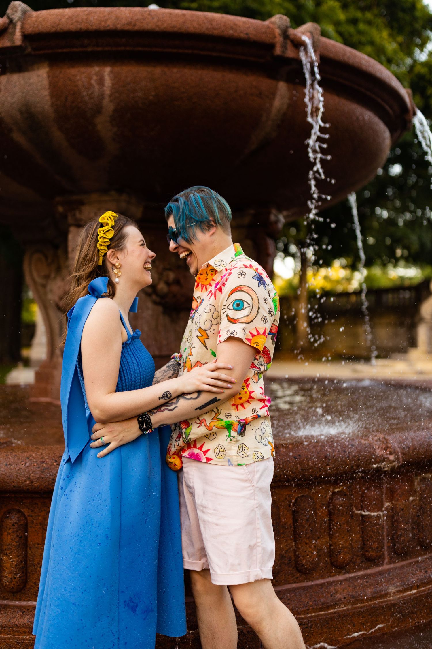 Joyful couple laughing together beside a rustic stone fountain in a park.