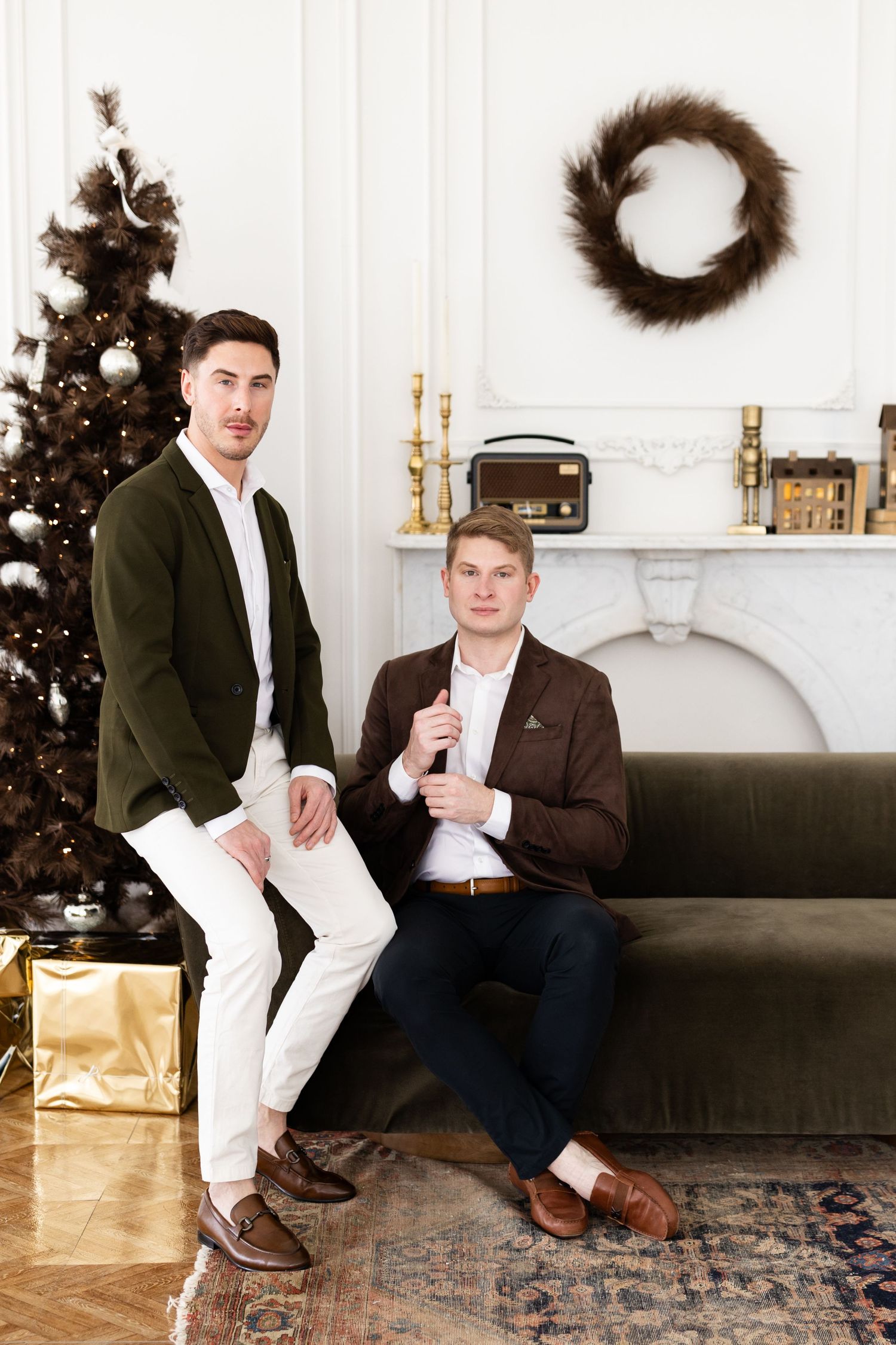 Two well-dressed men posing near Christmas tree and green velvet sofa in elegant holiday interior.
