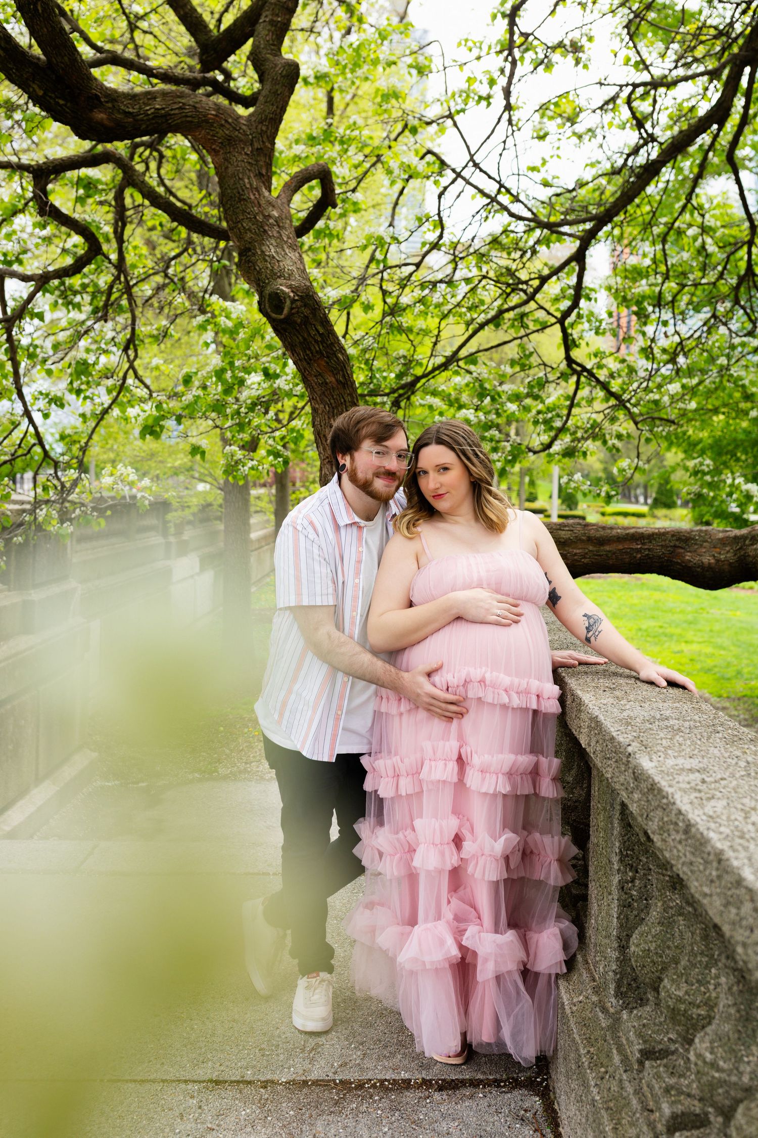 Couple embracing under blooming tree, woman in pink ruffled maternity gown leaning on stone wall.