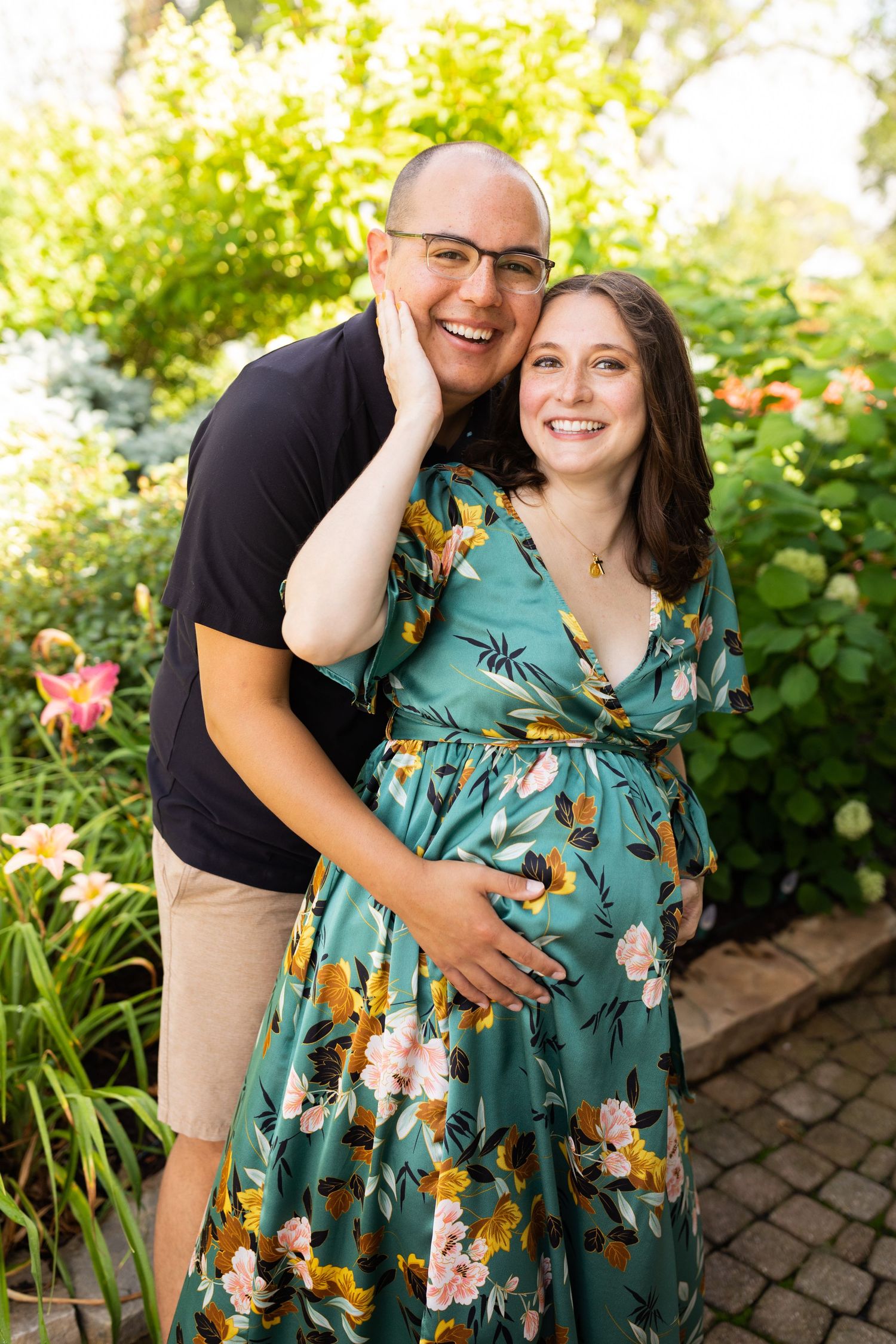Smiling pregnant couple posing in lush garden, woman in floral teal maternity dress.