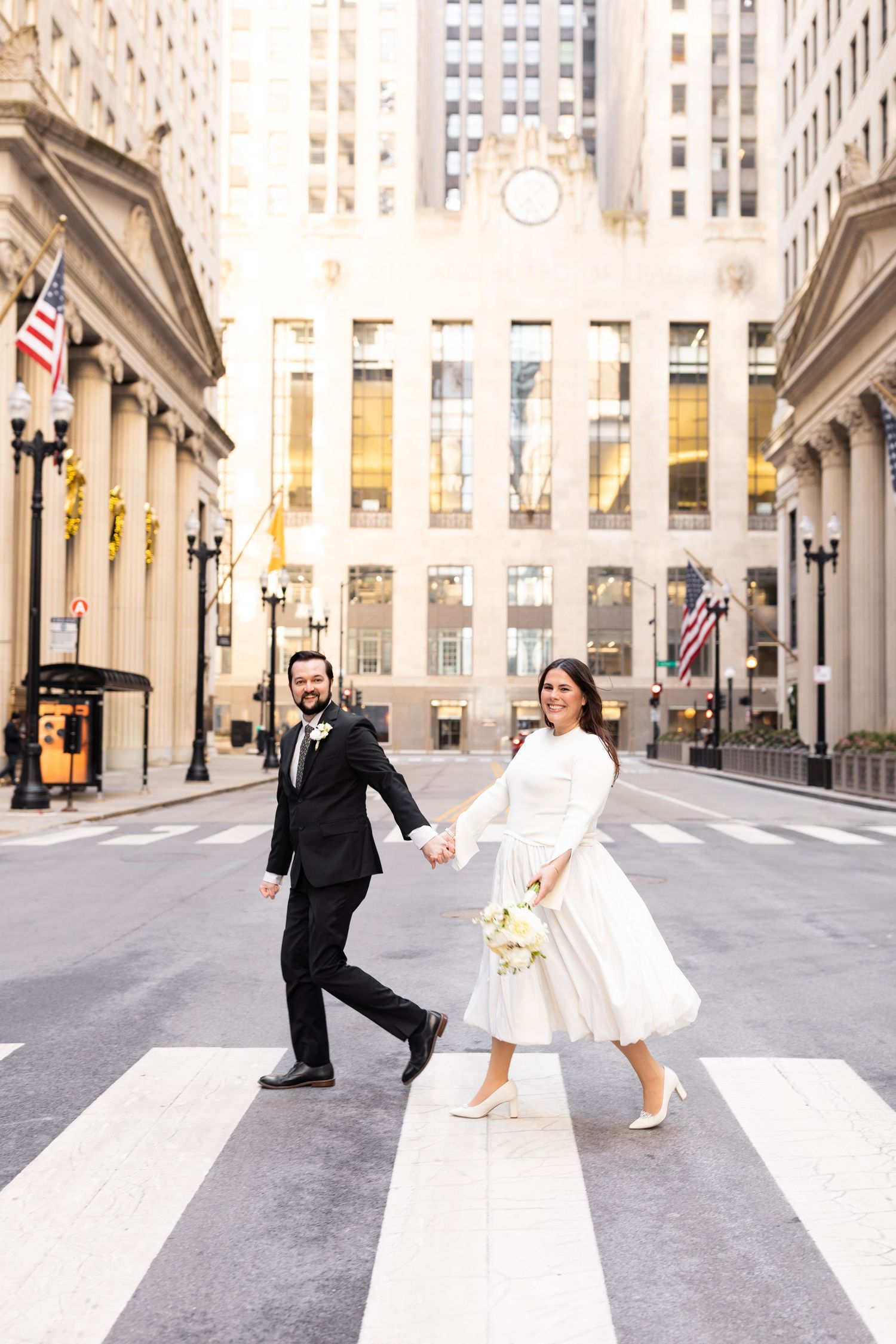 Joyful newlyweds crossing a downtown street holding hands in a sunlit city.