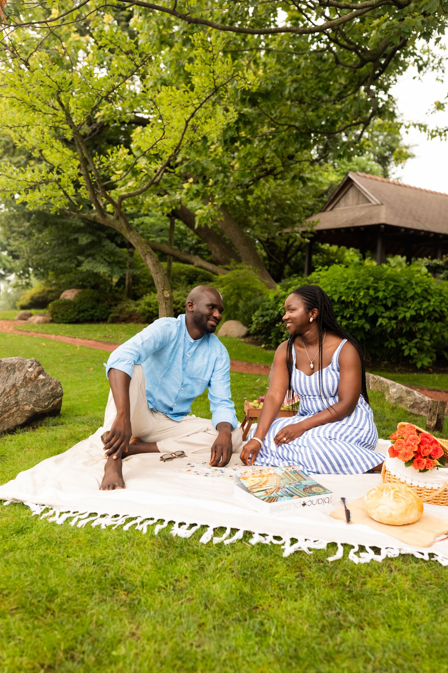 A happy couple sits on a blanket in a park, smiling at each other during a sunny outdoor picnic.