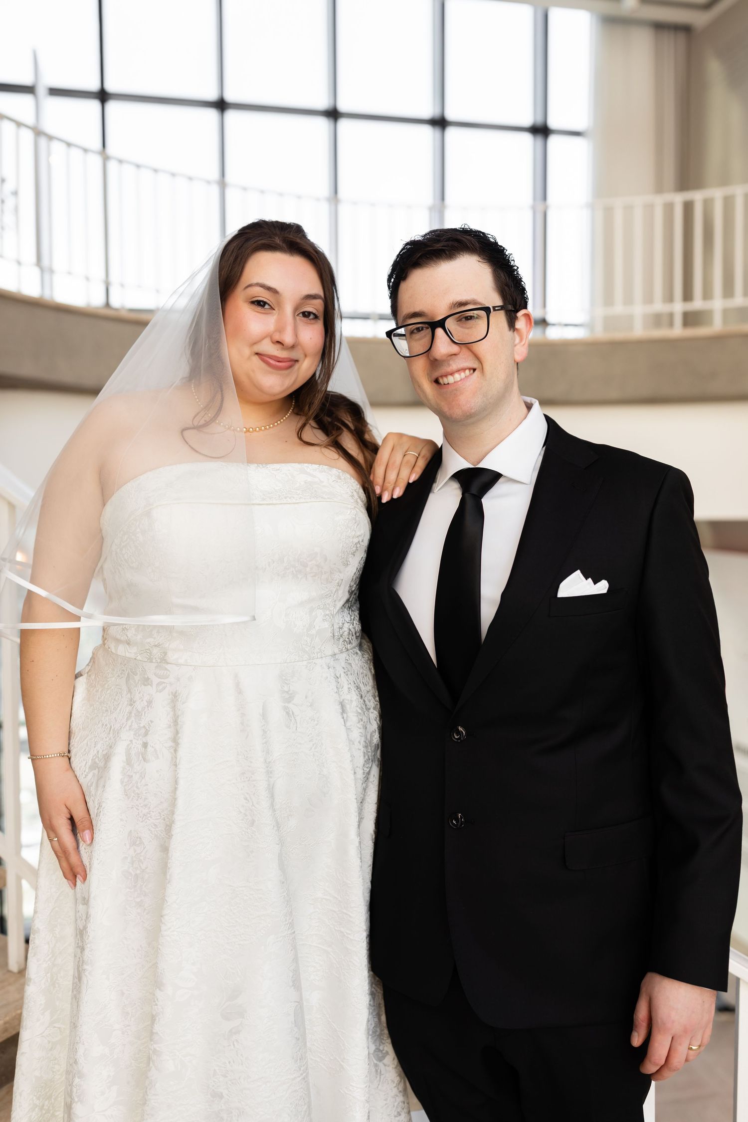 Smiling bride and groom posing together on staircase, she in white lace gown and veil, he in black suit.