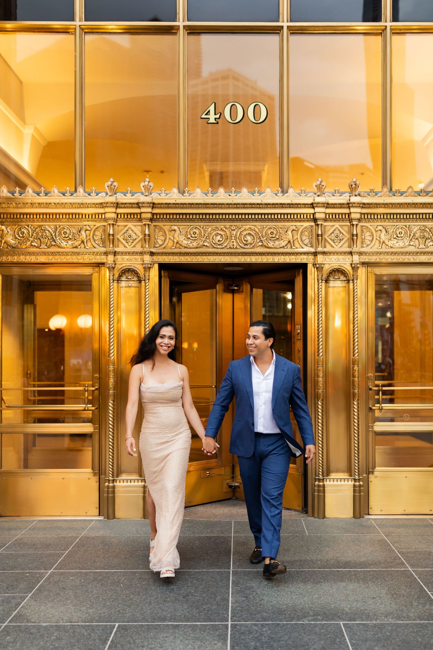 Smiling couple walking hand-in-hand out of grand gold-adorned 400 building entrance together.