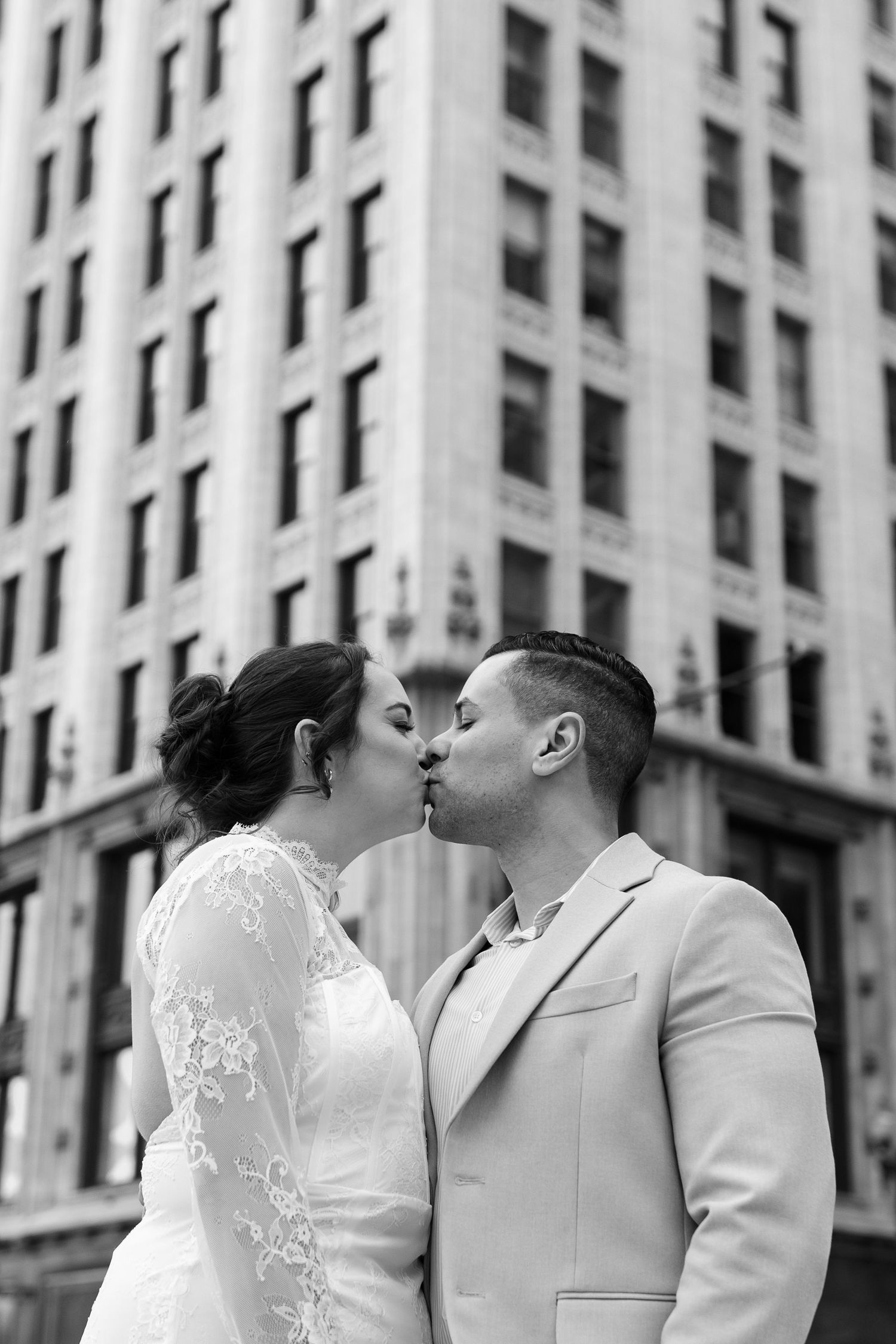 Bride and groom share a kiss on a city street with a tall building in the background, black and white photo.