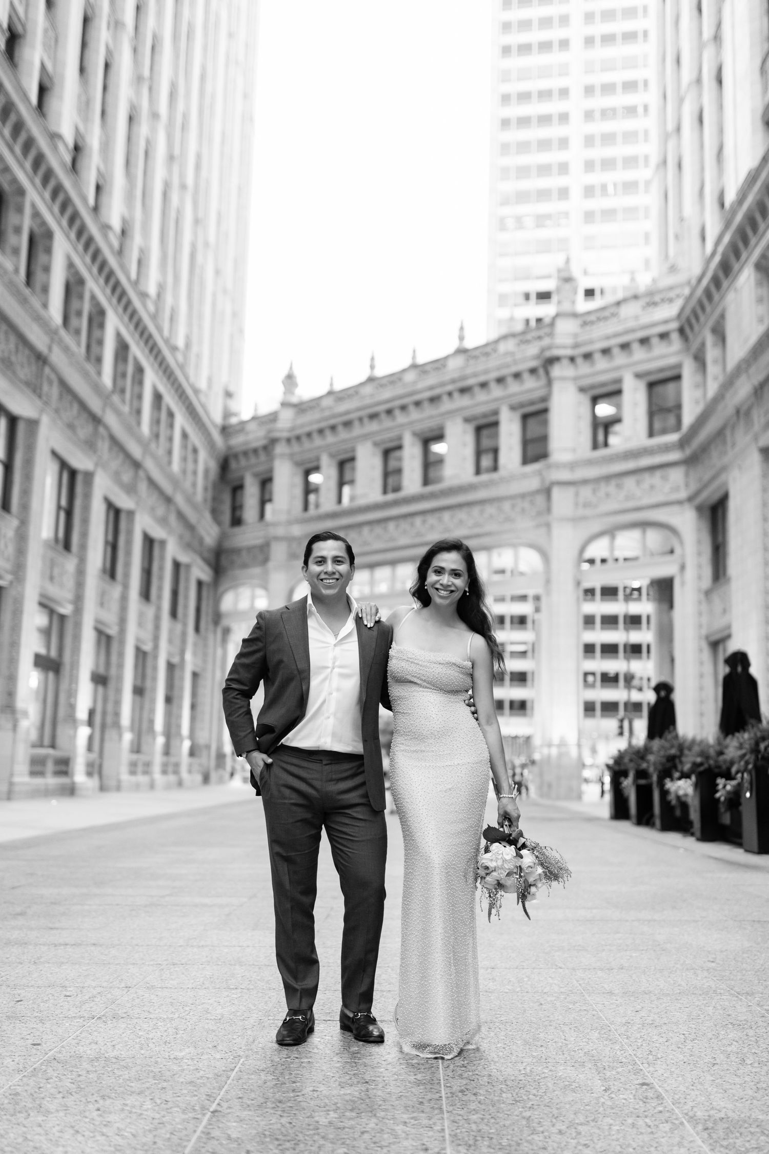 Black and white full-length portrait of engaged couple standing before ornate city building facade.