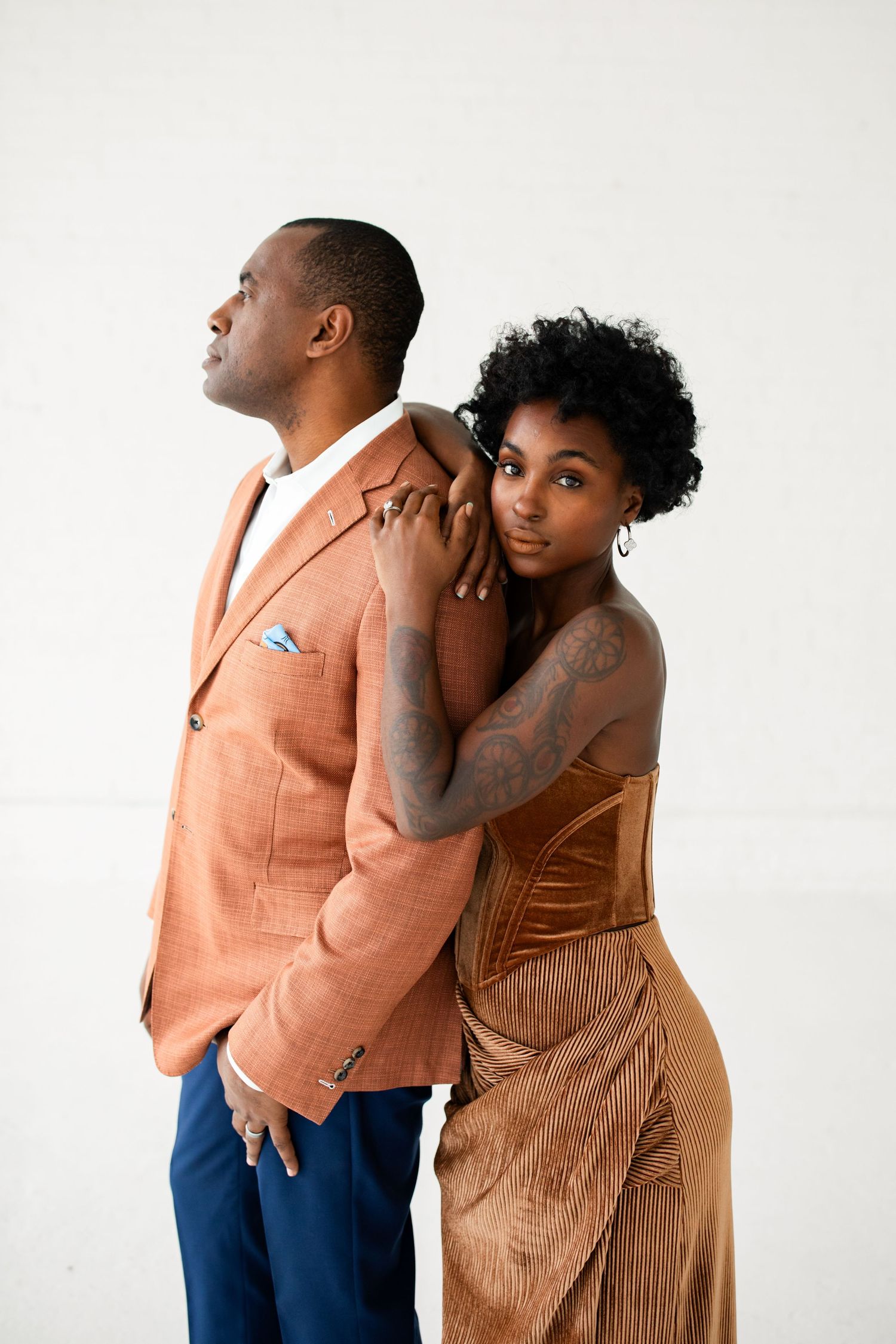 Elegant couple posing together, woman in gold velvet dress leaning on man in rust blazer against white background.