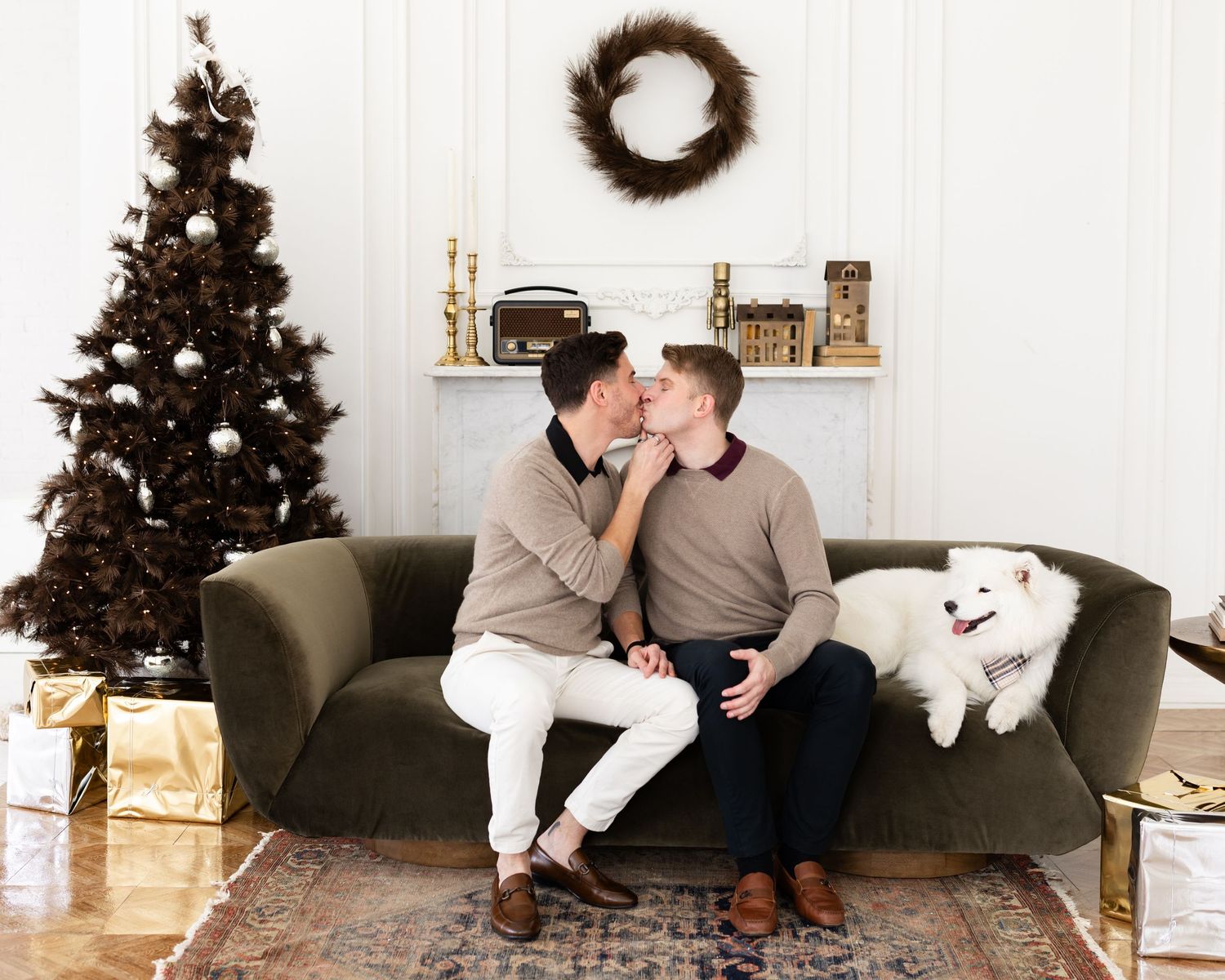 Two men sharing a kiss on green velvet sofa with white Samoyed dog in Christmas decorated room.