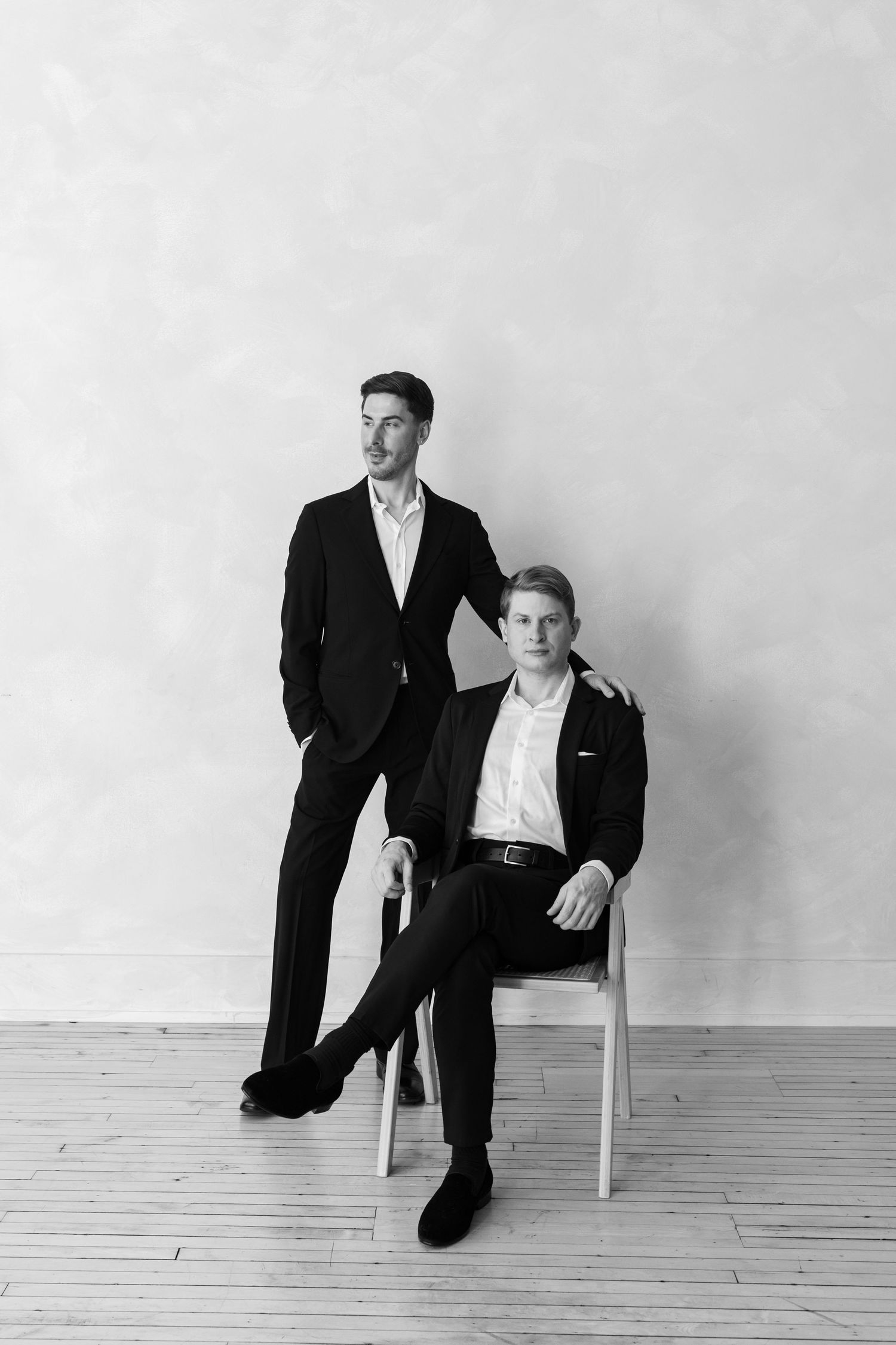 Black and white portrait of two men in dark suits, one standing and one seated on a chair in a studio.