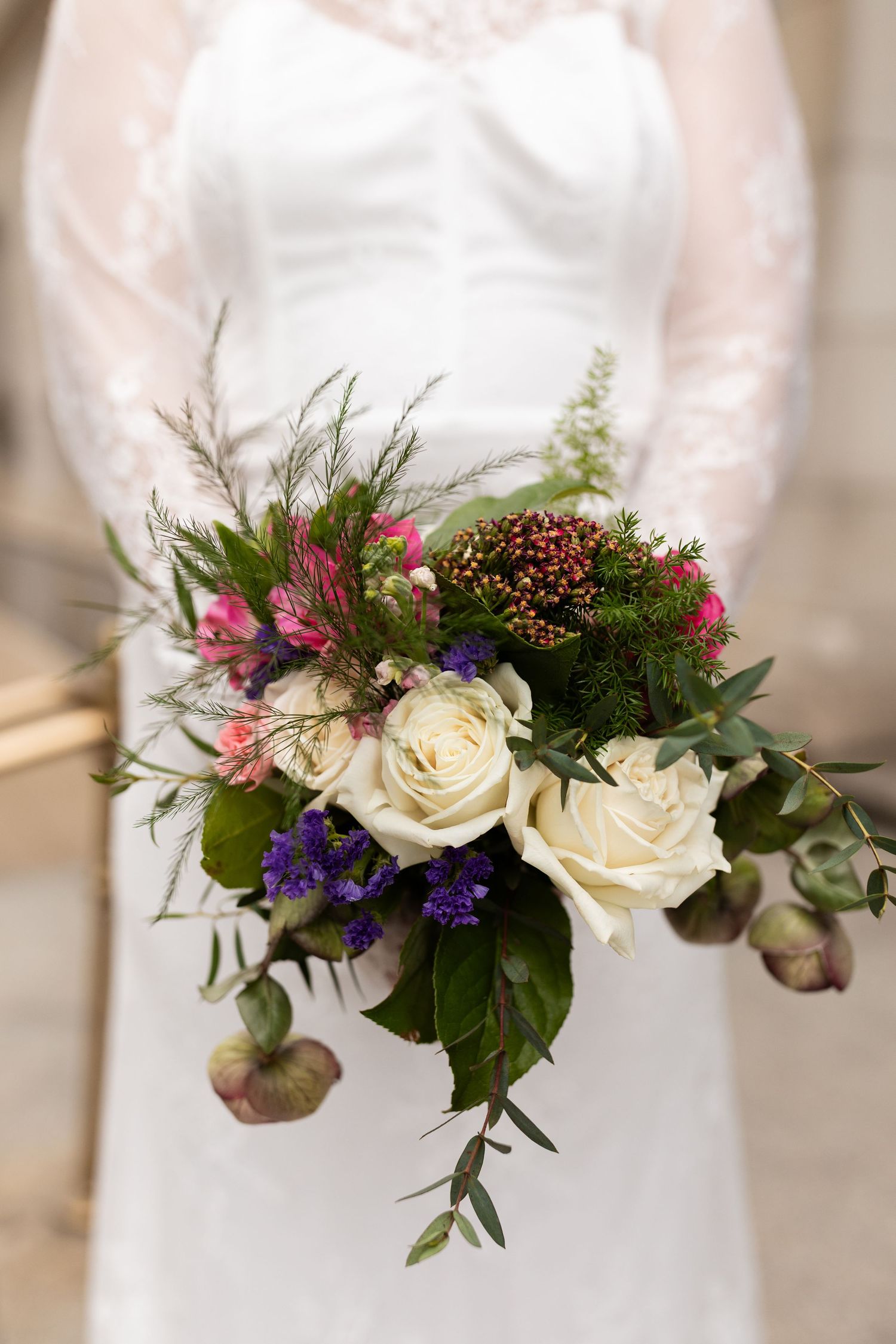 Close-up of bridal bouquet featuring white roses, purple statice, and pink flowers held by lace-gowned bride.