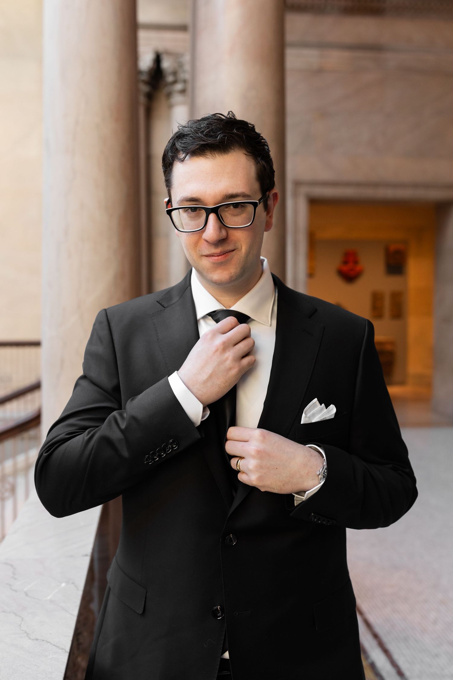 Dapper groom in black suit and glasses adjusting his tie beside classical stone columns.