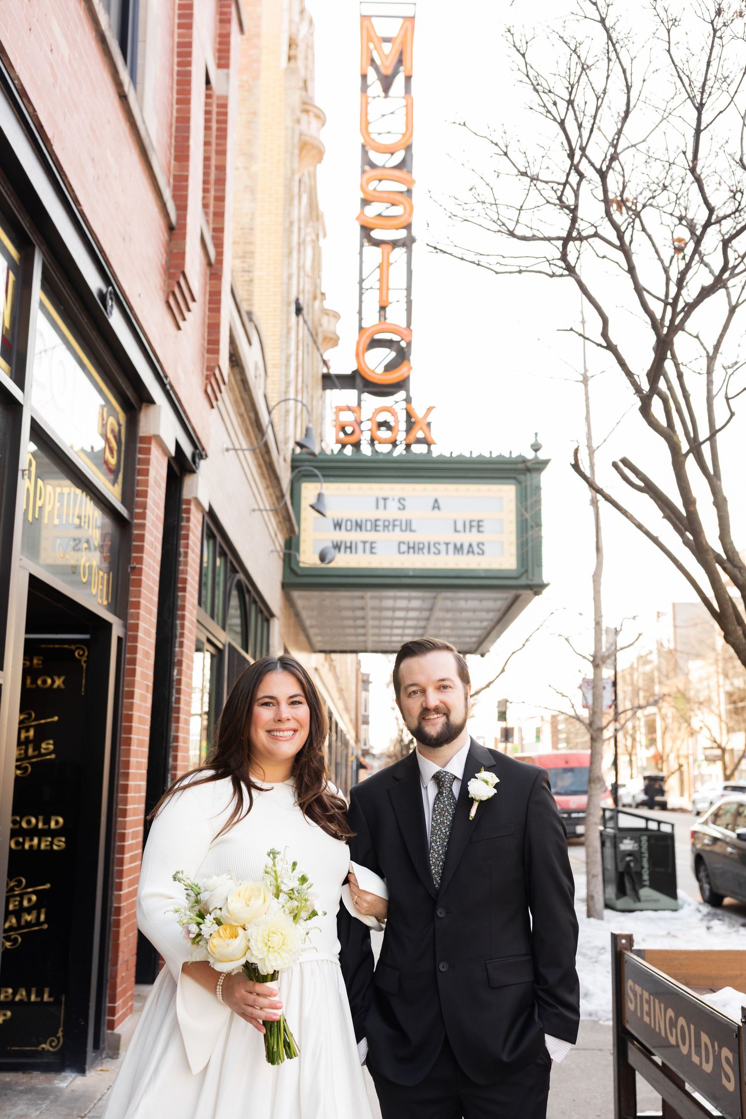 Smiling bride holding white bouquet and groom posing together outside Music Box Theatre in Chicago winter wedding.