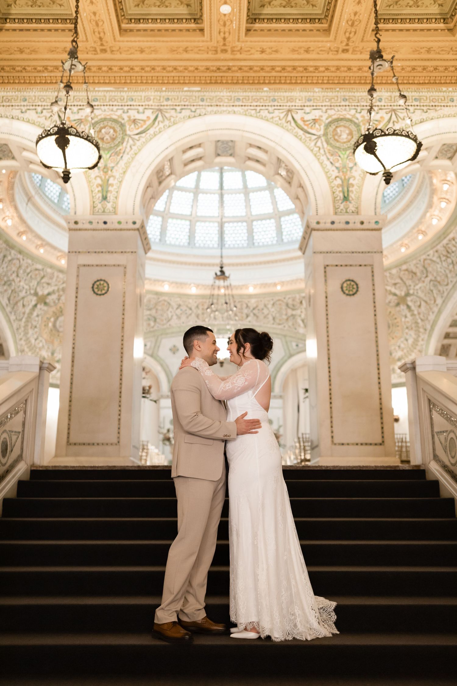 Bride and groom embrace and nearly kiss on a grand staircase inside an ornate historic Chicago building.