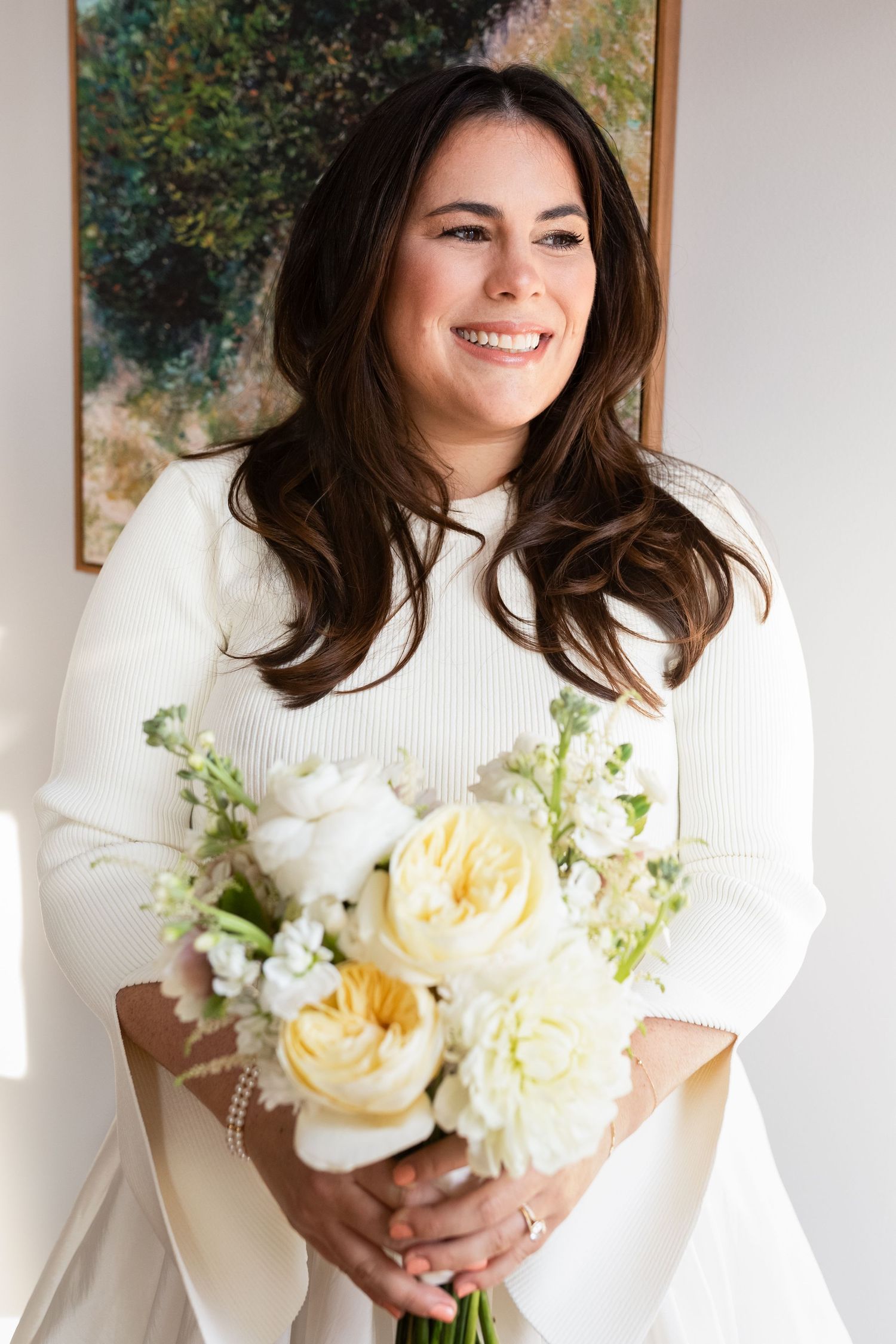 Smiling dark-haired bride in white dress holds yellow and white floral bouquet indoors.