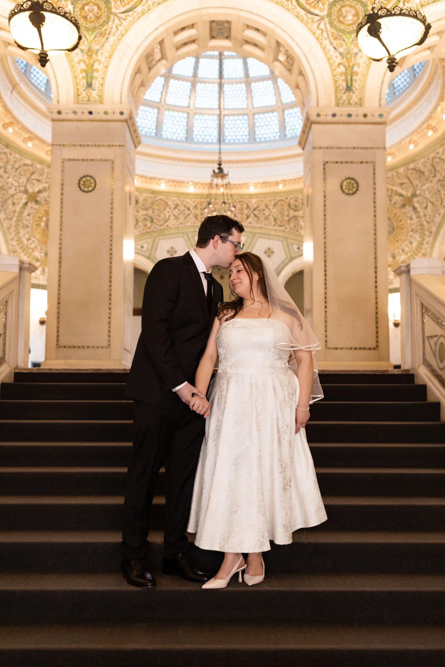 Couple shares romantic moment on grand staircase beneath gilded dome of historic building.