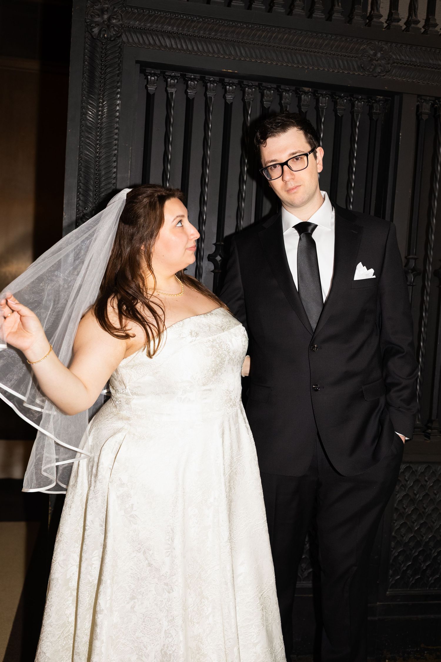 Bride in strapless white gown holds veil beside groom in black suit before ornate dark backdrop.