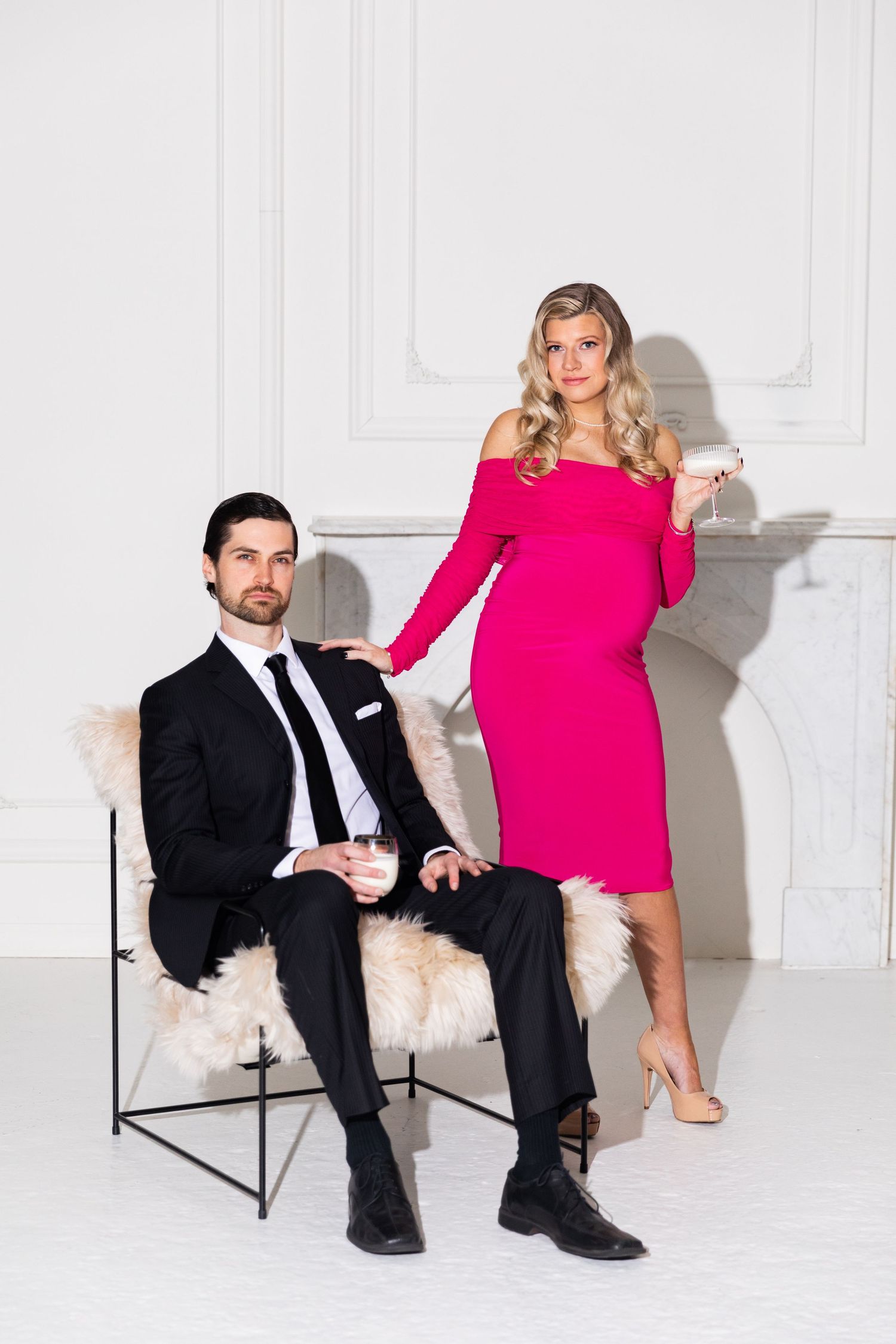 Elegant pregnant couple in formal attire, woman in pink off-shoulder dress holding martini glass.