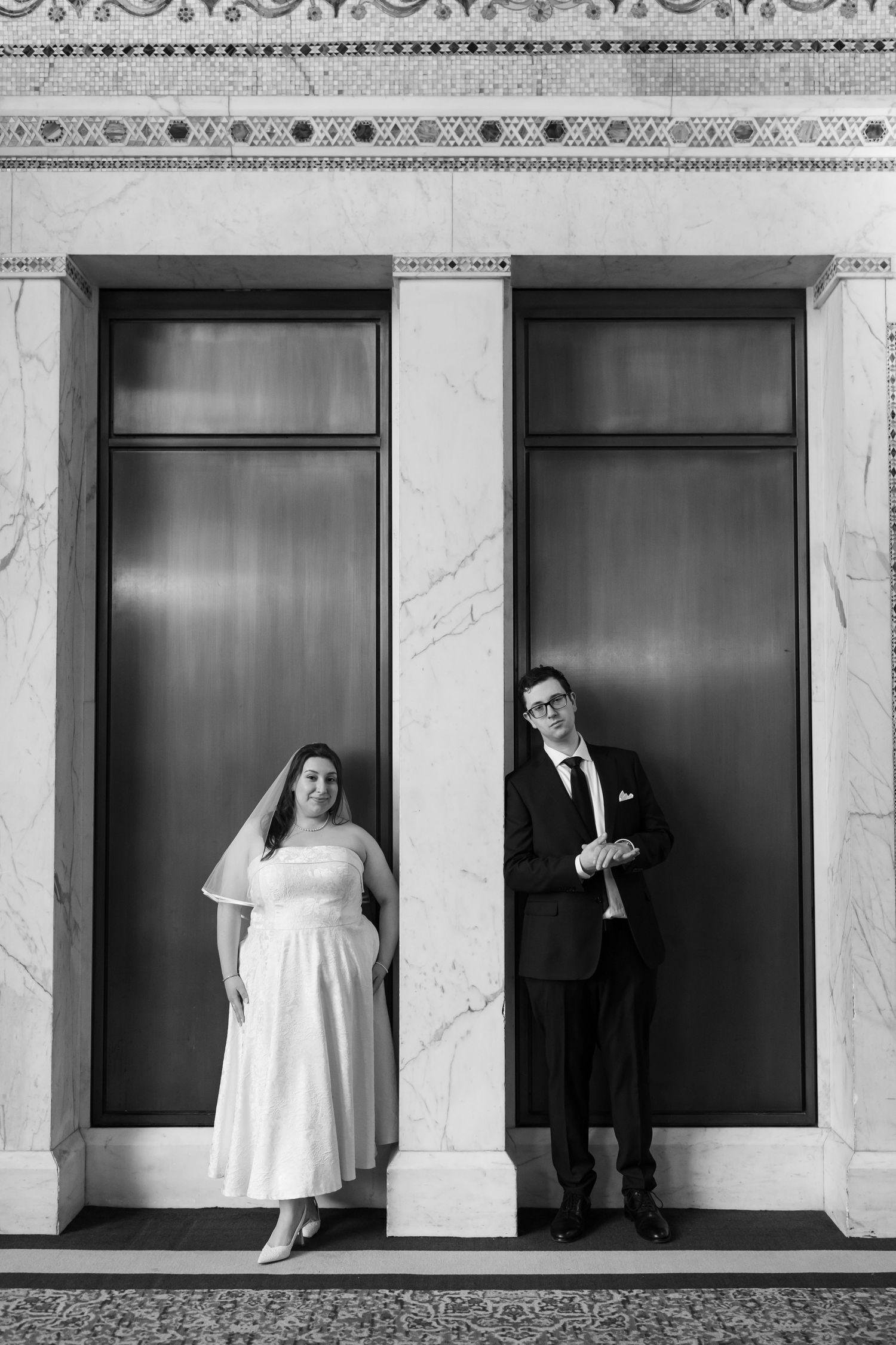 Black and white photo of bride and groom standing apart between marble columns and tall doors.