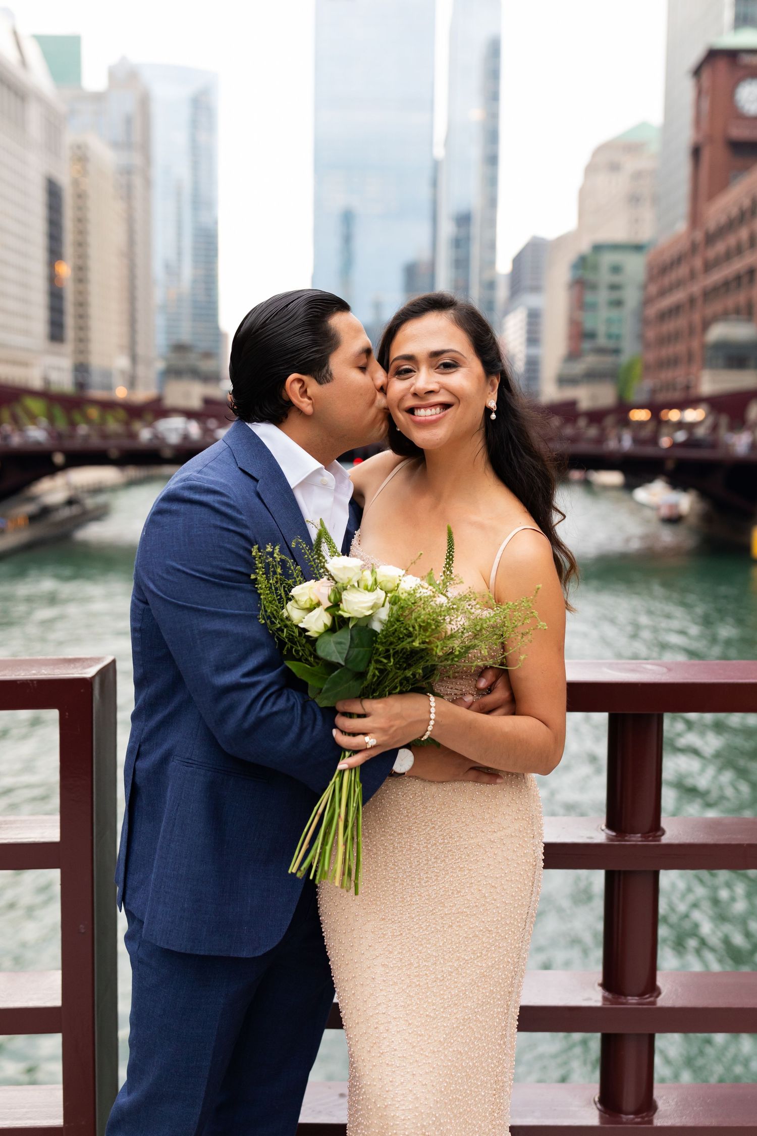 Groom kissing bride on cheek while she smiles holding white rose bouquet on Chicago Riverwalk.