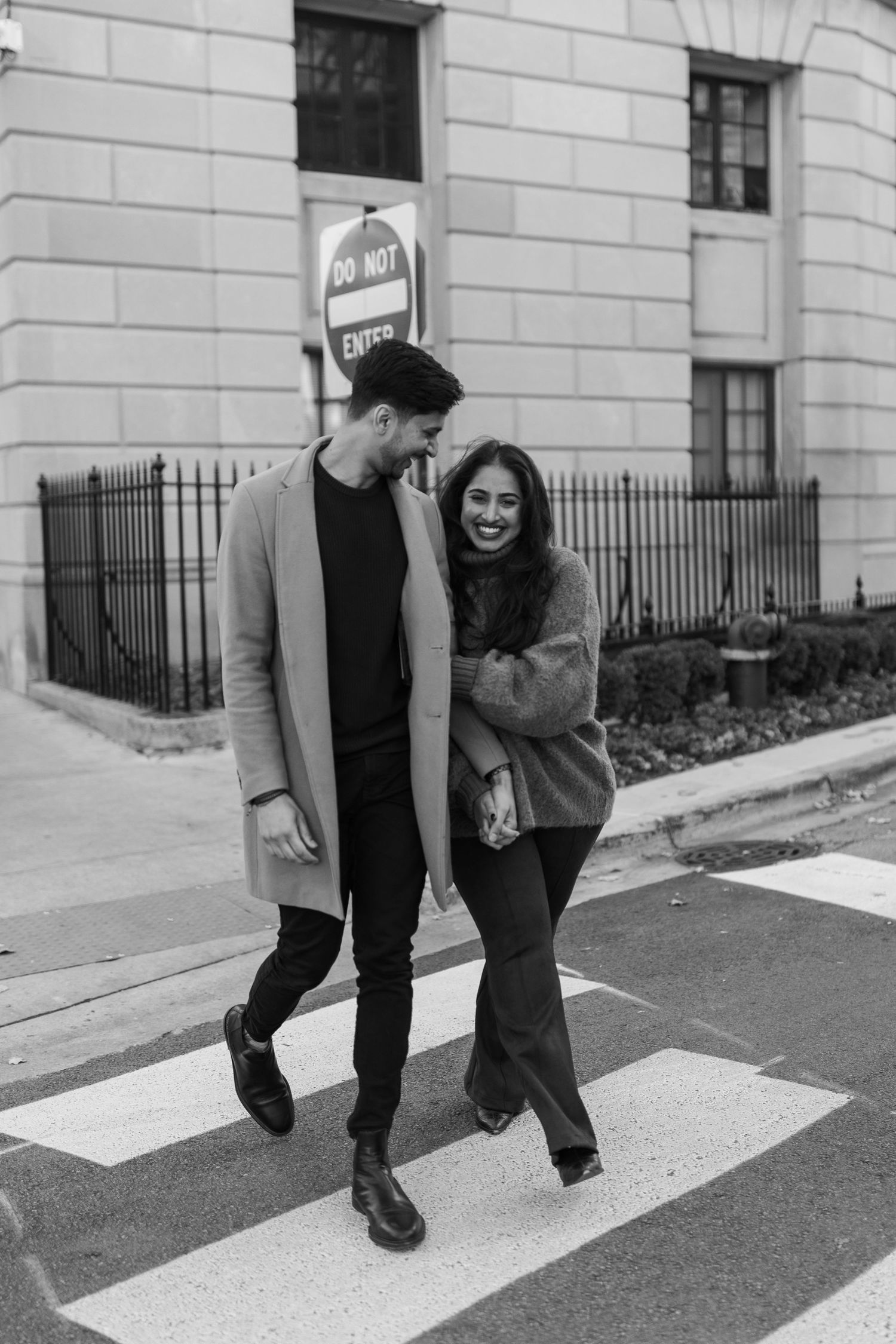 Black and white photo of laughing couple crossing street, man in long coat, woman in fuzzy sweater holding hands.