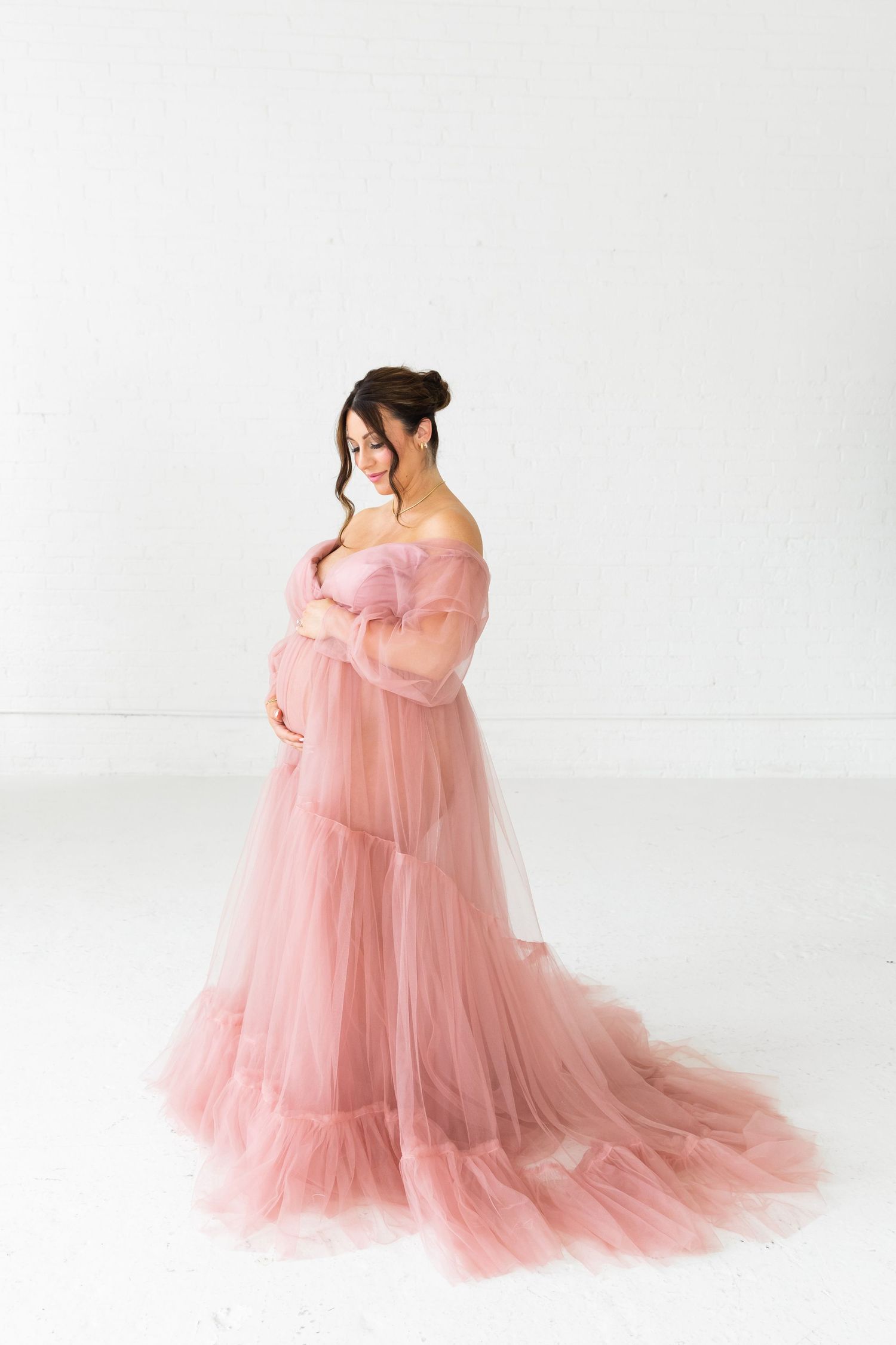 Pregnant woman in elegant pink tulle off-shoulder gown cradling baby bump against white background.