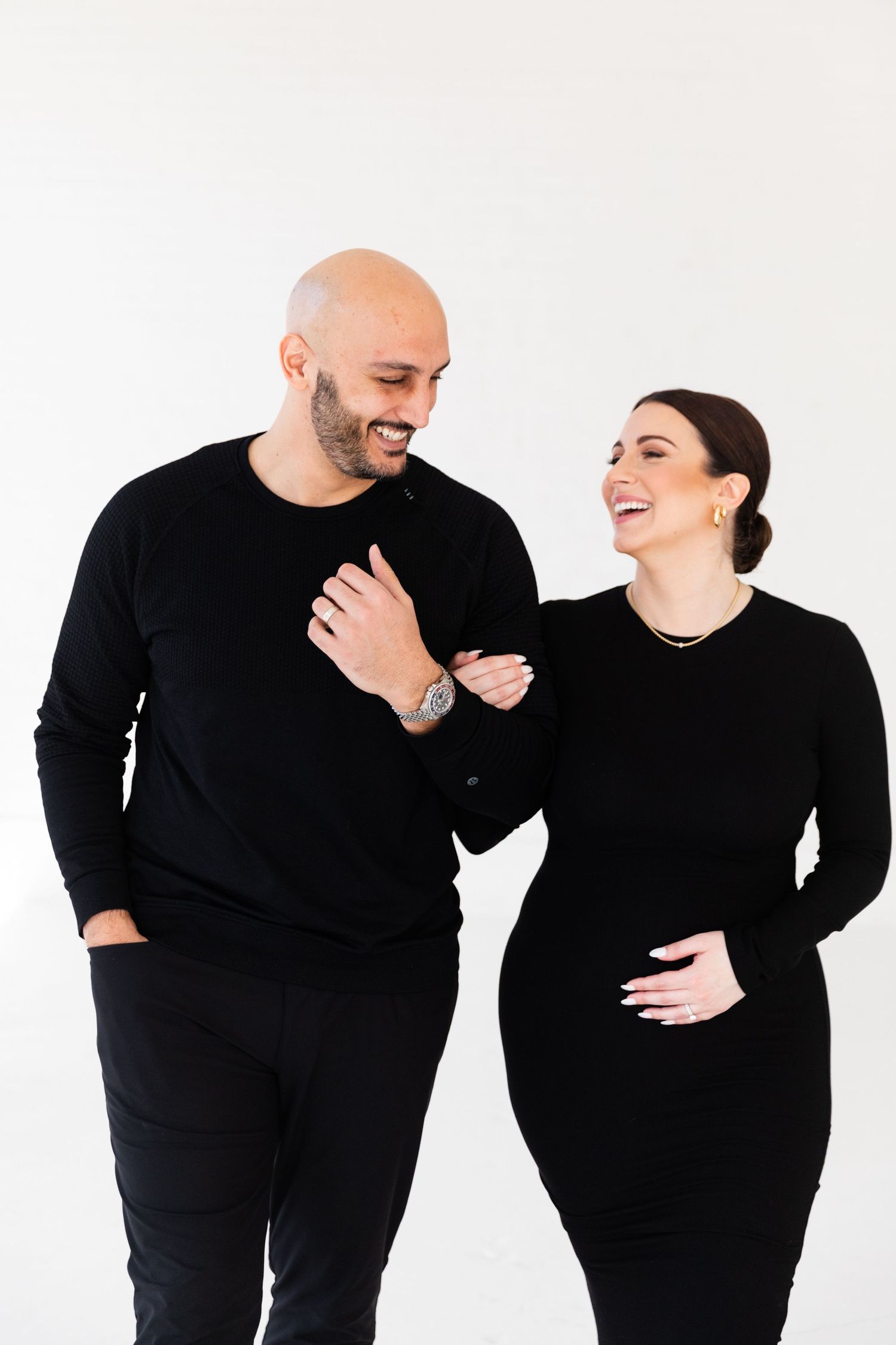 Happy expecting couple in matching black outfits laughing together against white background.