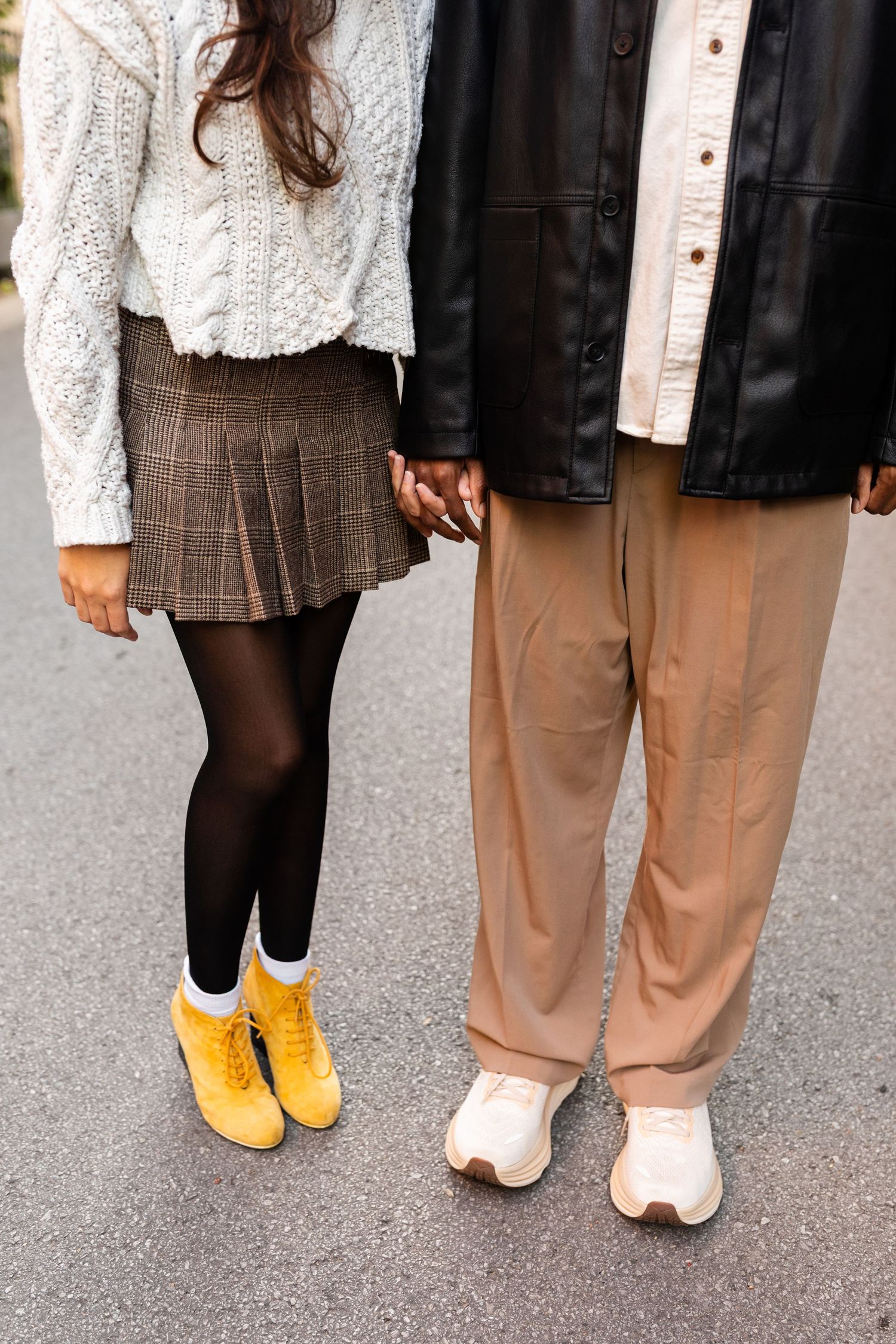 Close-up of couple holding hands, woman wearing yellow boots and plaid skirt, man in beige wide-leg pants.