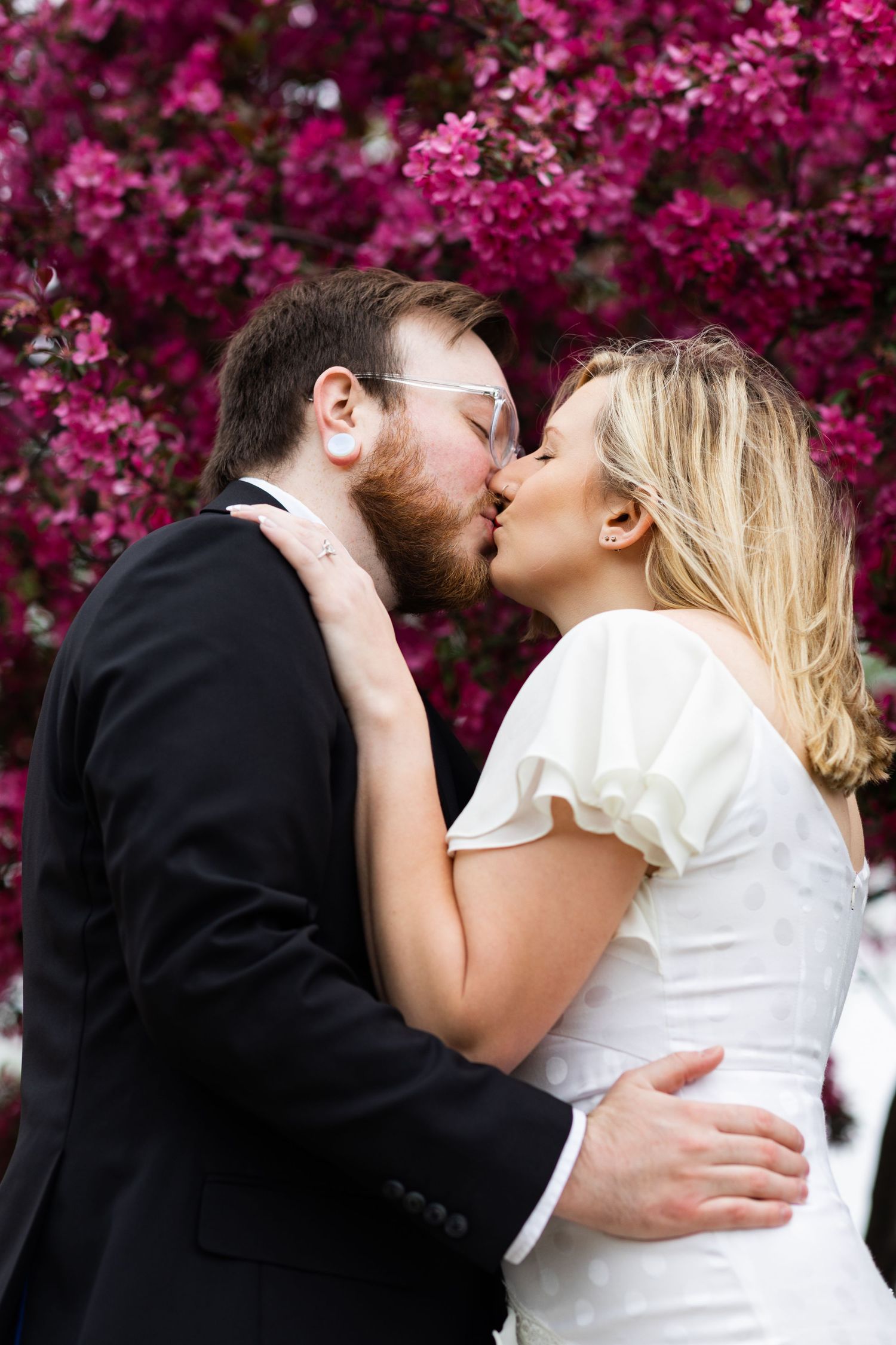 Bride and groom share a romantic kiss in front of stunning pink blooming cherry blossom trees.