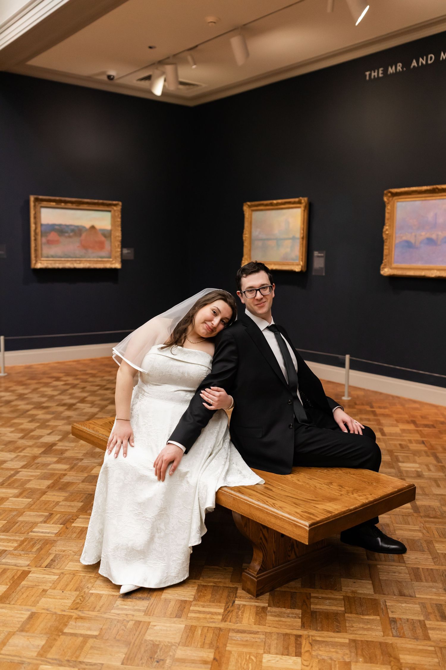 Bride and groom posing on a wooden bench in a Monet gallery, leaning together in wedding attire.