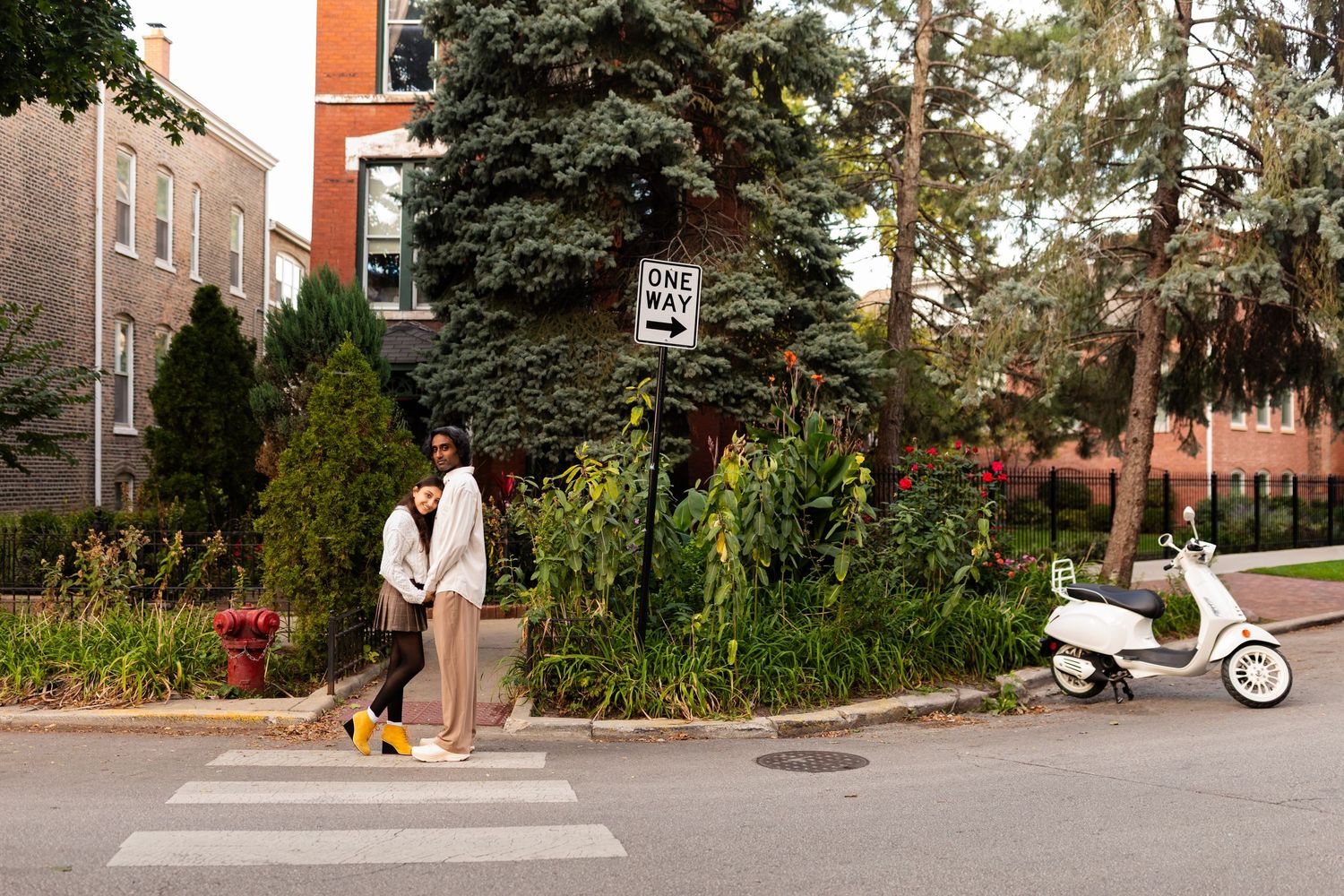 Couple standing at a crosswalk near a one-way sign with a white scooter and brick buildings in background.