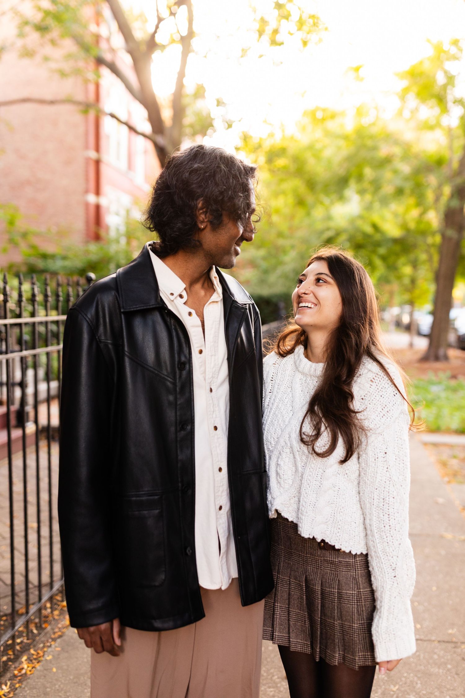 Happy interracial couple laughing together on sunny autumn sidewalk, man in black leather jacket with arm around woman.