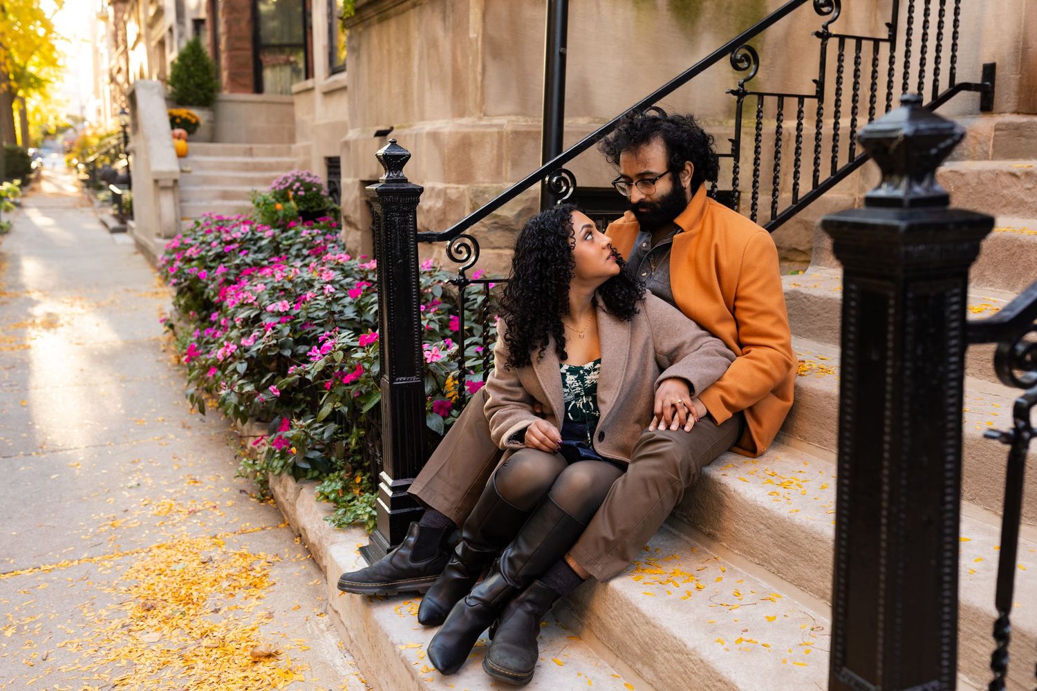 A couple sits together on sunlit steps surrounded by colorful flowers and autumn foliage.
