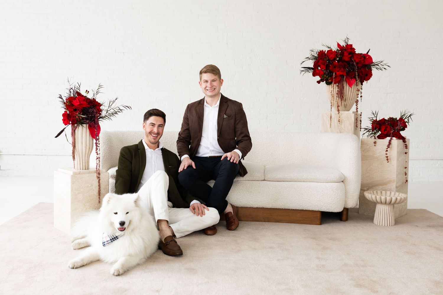 Two grooms smile together on white sofa with Samoyed dog and elegant red floral arrangements nearby.