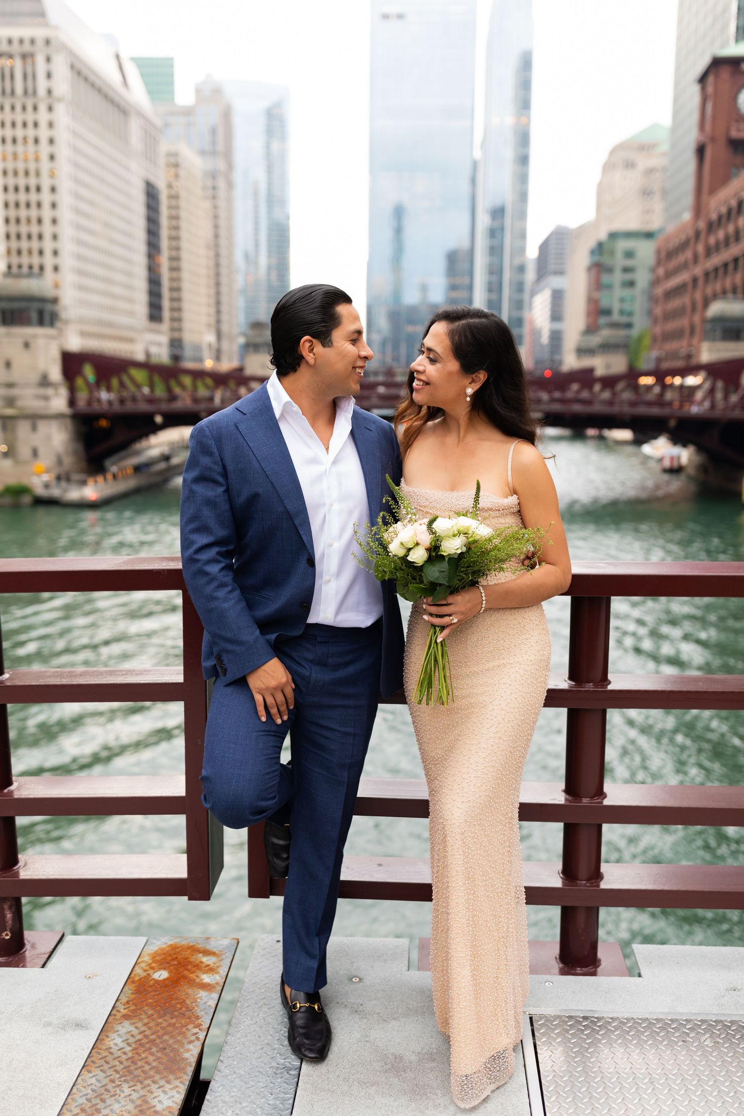 Couple in wedding attire poses on a bridge over the Chicago River with city skyline in the background.