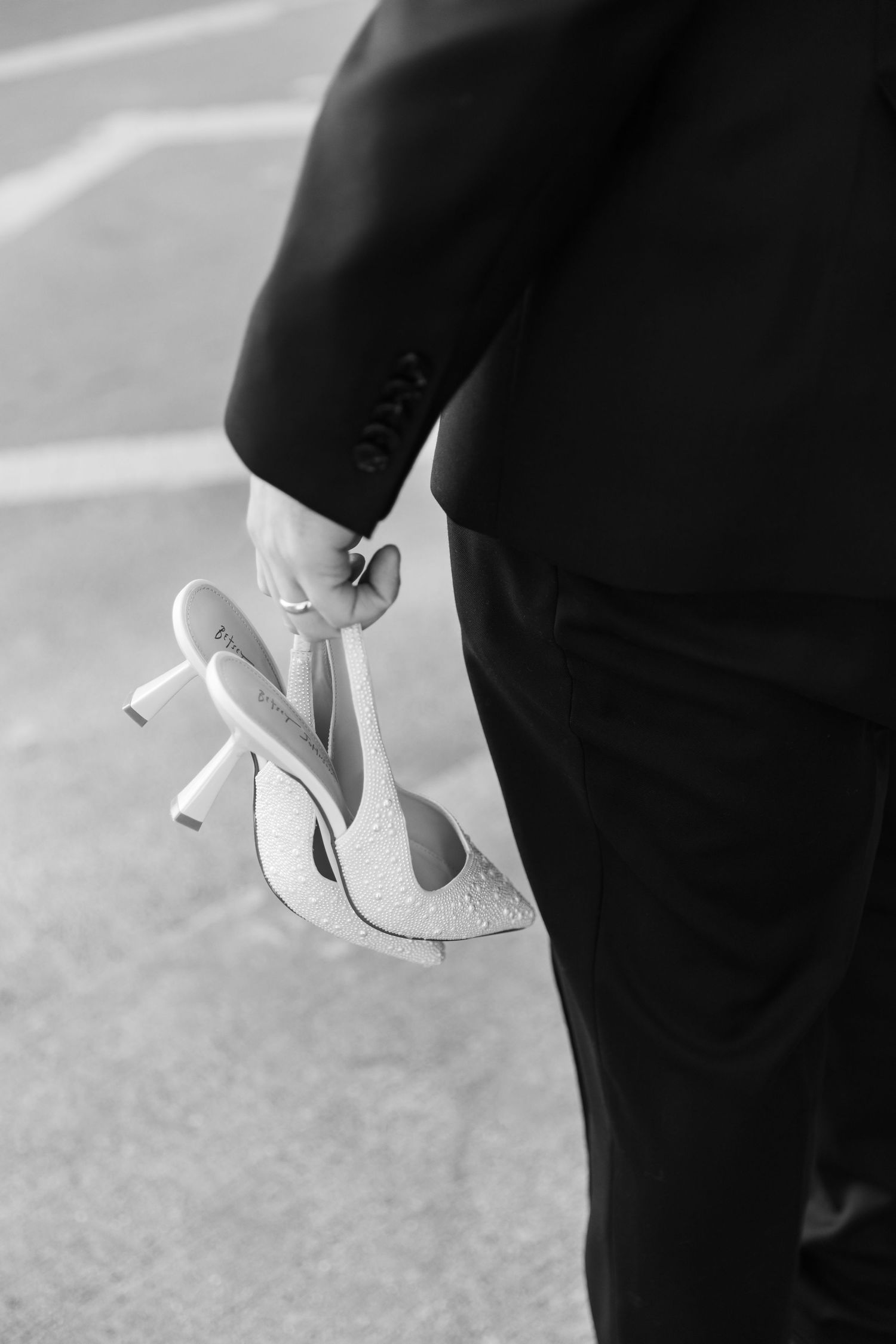 Black and white image of groom in dark suit carrying sparkly bridal heels at outdoor wedding.