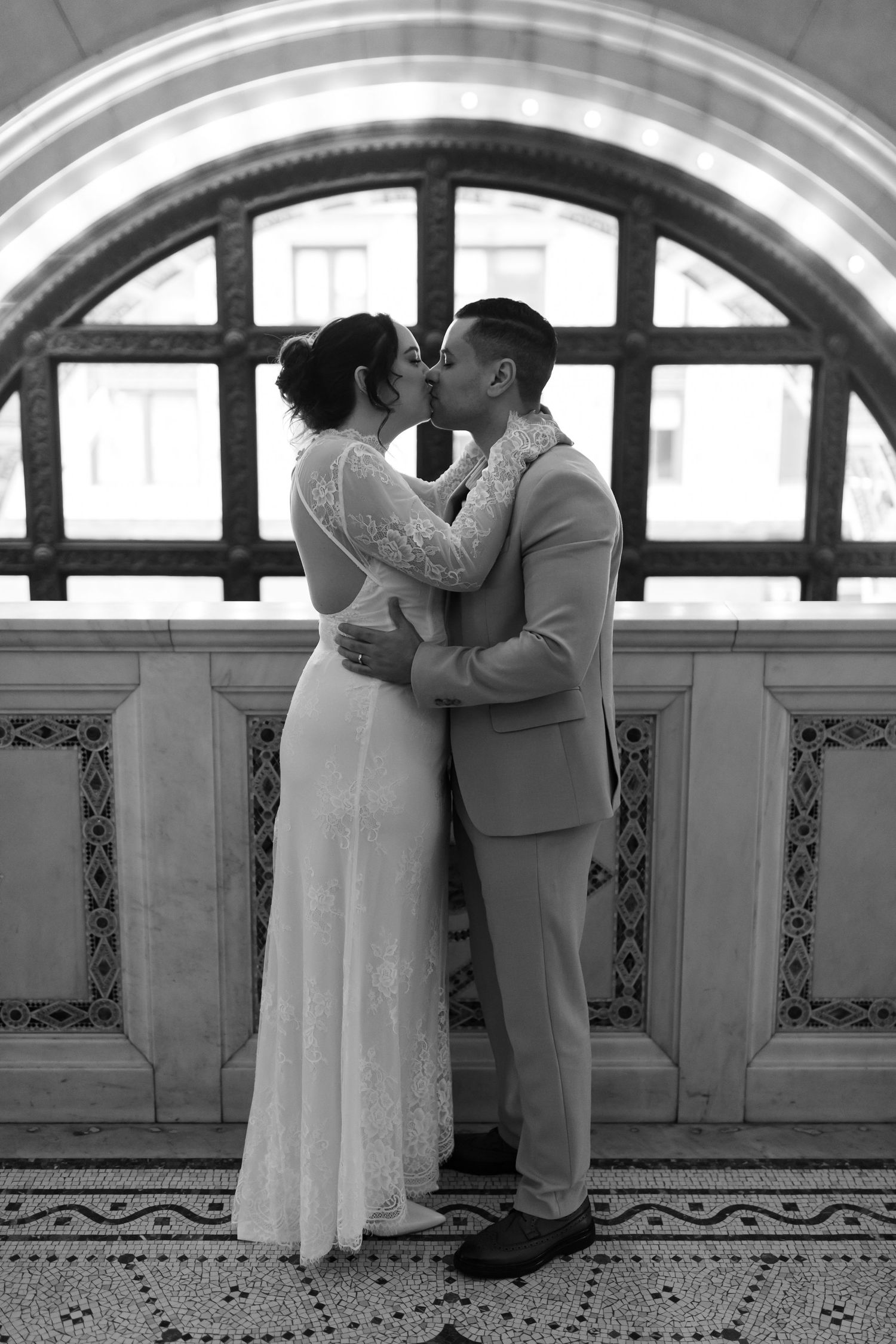 Black and white photo of a bride and groom kissing in front of an arched window in a grand hall.
