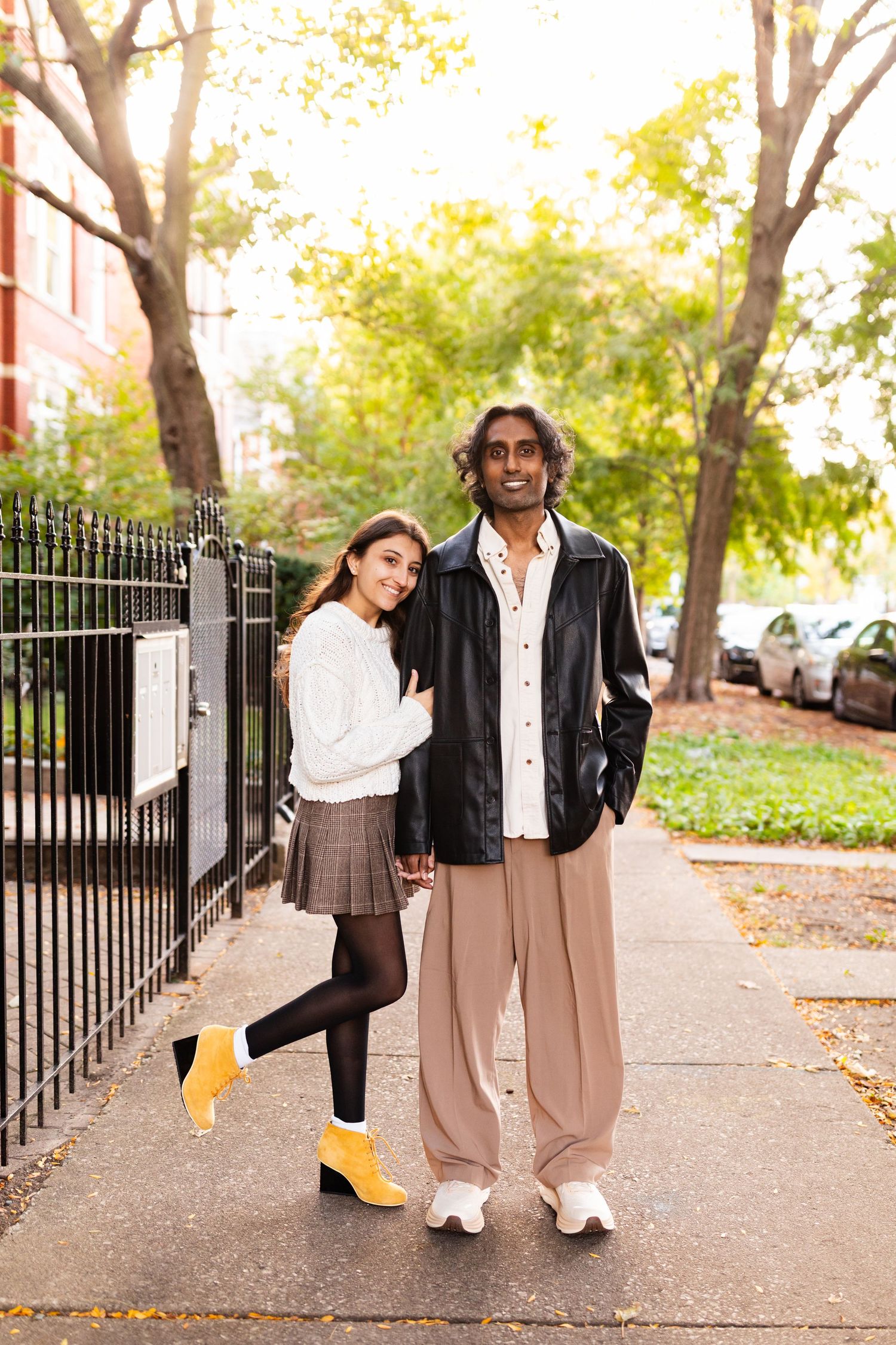 Interracial couple holding hands on a tree-lined sidewalk, woman in white sweater and yellow boots, man in black leather jacket.