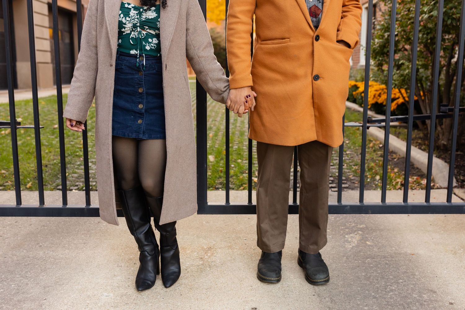 A couple holds hands in front of a black iron fence, dressed in coordinating fall outfits.