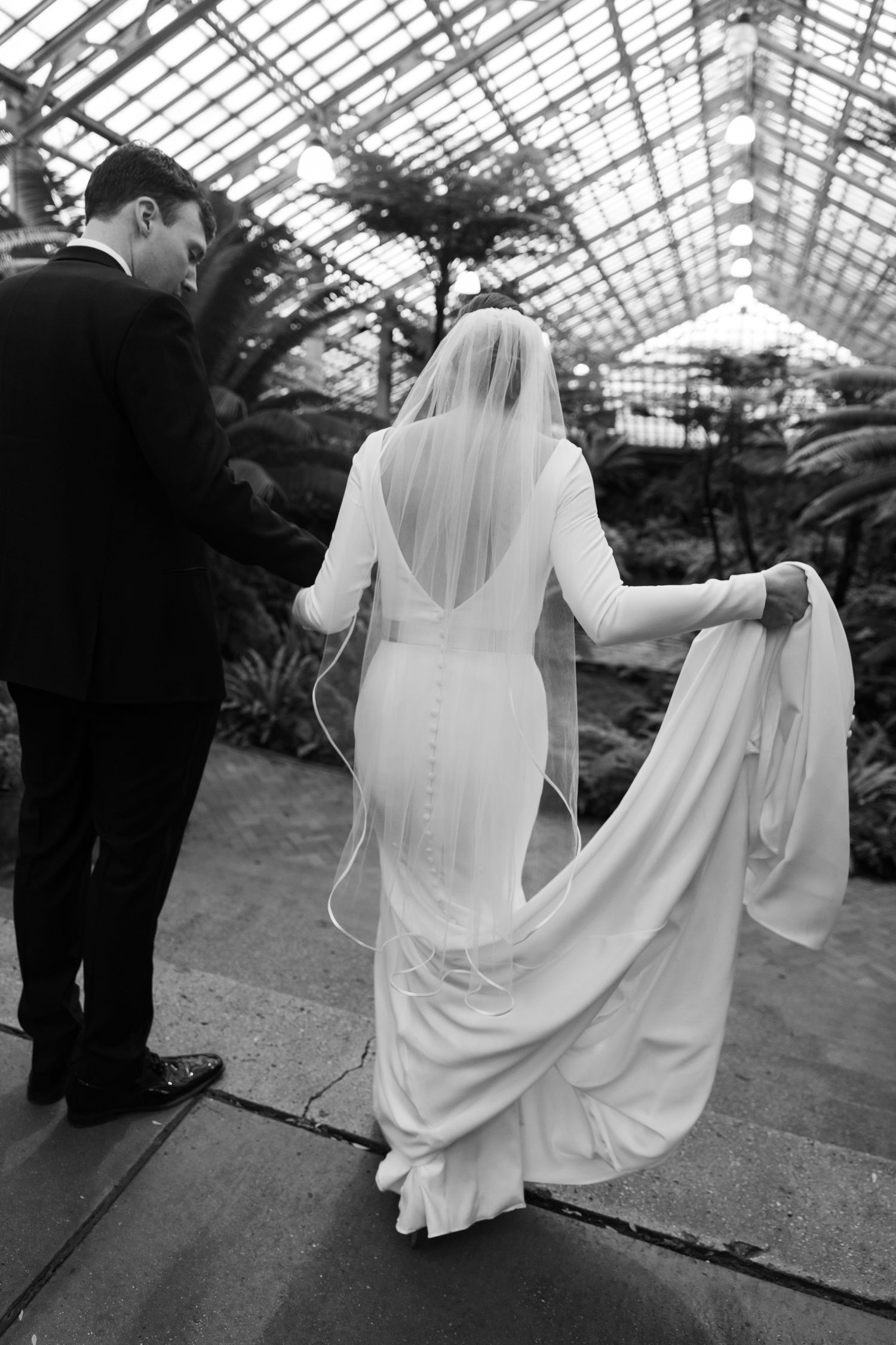 Black and white photo of a bride and groom walking through a lush greenhouse conservatory.