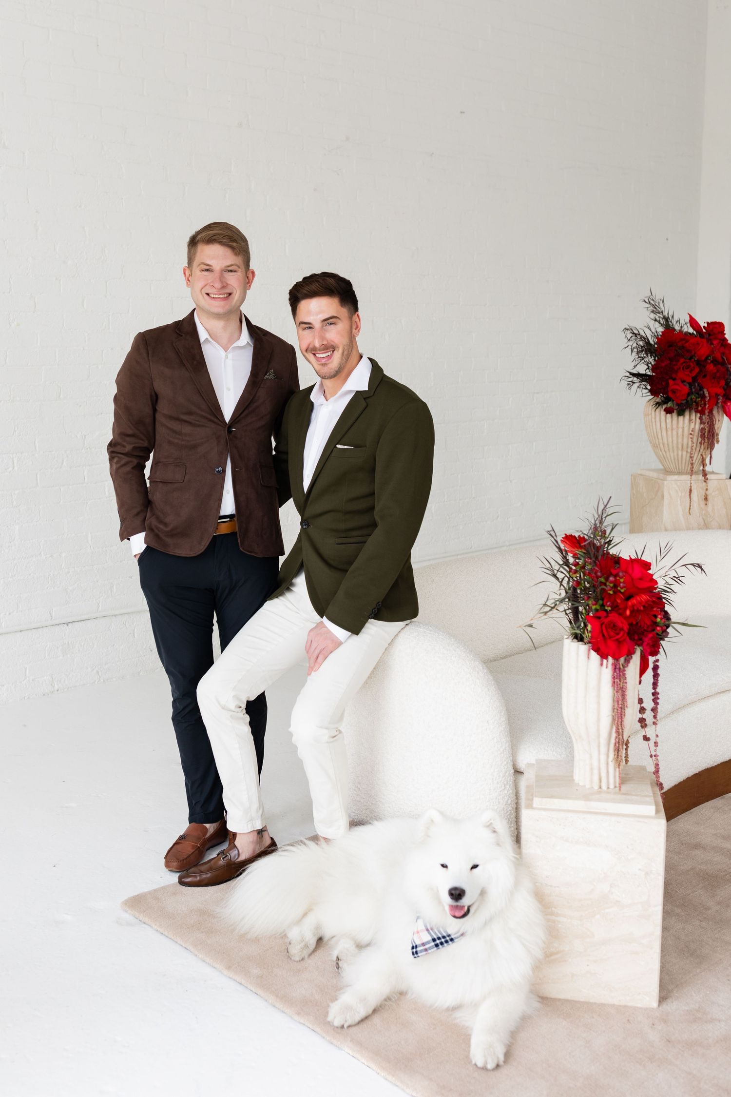 Two men in blazers pose with fluffy white Samoyed dog beside holiday floral arrangements.