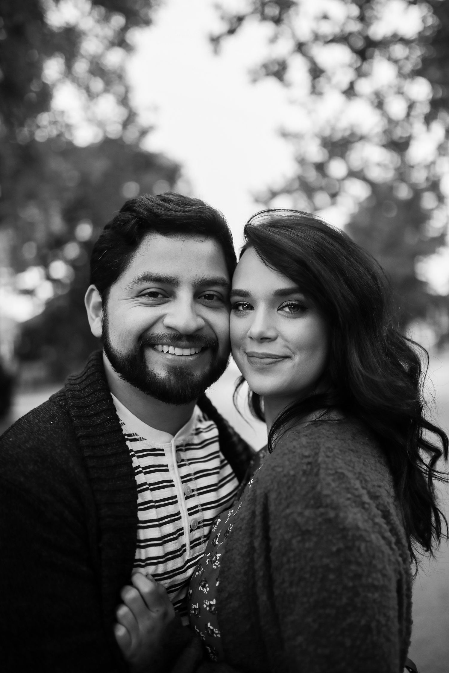 Black and white portrait of smiling Hispanic couple embracing outdoors with soft bokeh tree background.
