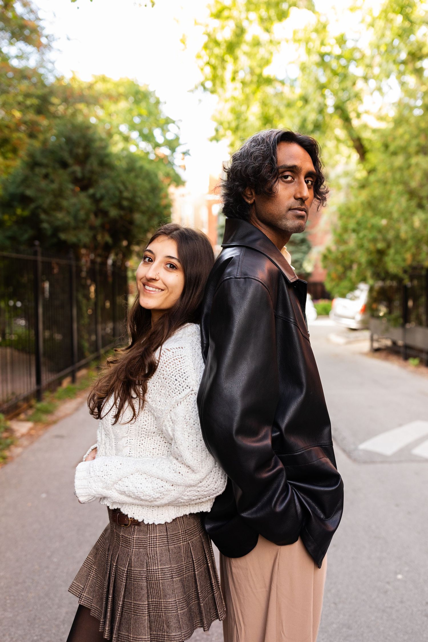 Smiling young woman and man standing back to back on a green tree-lined city street.
